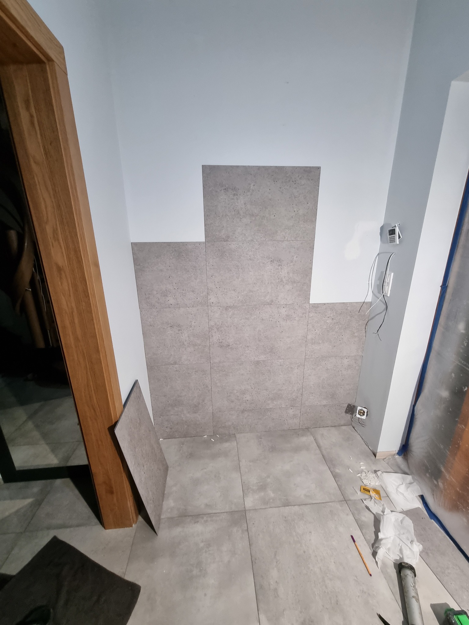 Fragment ściany łazienki w trakcie układania szarych, kwadratowych płytek ceramicznych, widoczne wystające przewody elektryczne i narzędzia na podłodze.