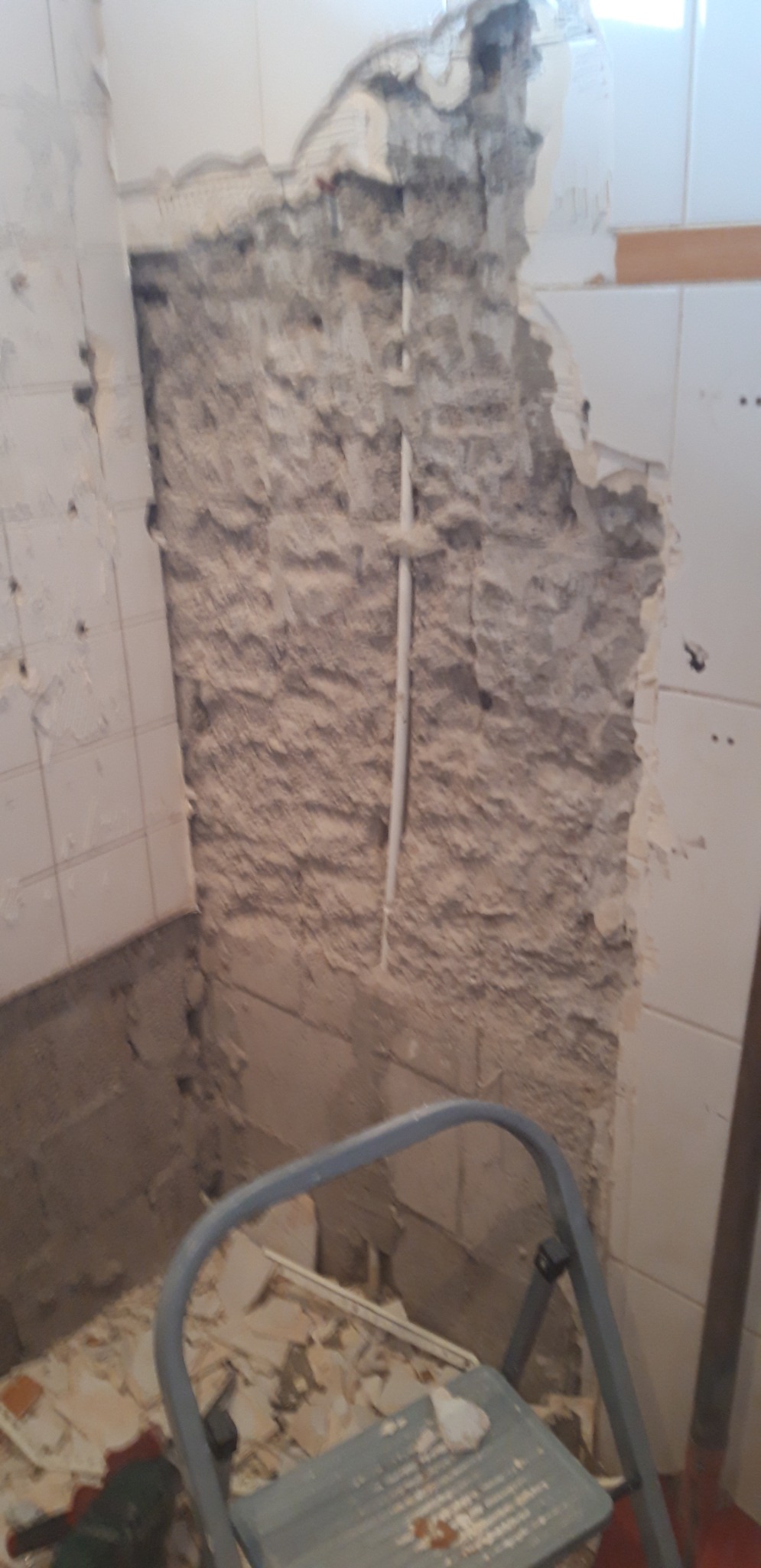 Demontaż starych płytek w łazience, odsłonięty surowy mur z wystającą rurą, widoczny fragment drabiny malarskiej i gruz na podłodze.