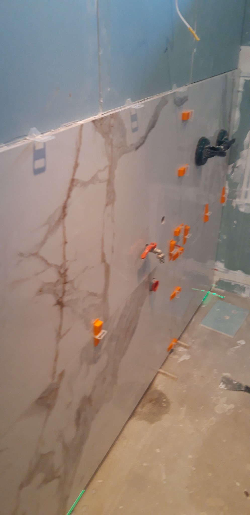 Układanie płytek imitujących marmur na ścianie w łazience, widoczne krzyżyki dystansowe, poziomica laserowa i fragment instalacji wodnej.