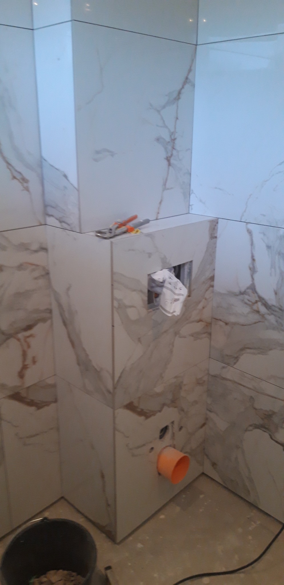 Łazienka w trakcie wykańczania: obudowa stelaża WC z płytkami imitującymi marmur, widoczne narzędzia, rura kanalizacyjna i otwór na przycisk spłukujący.