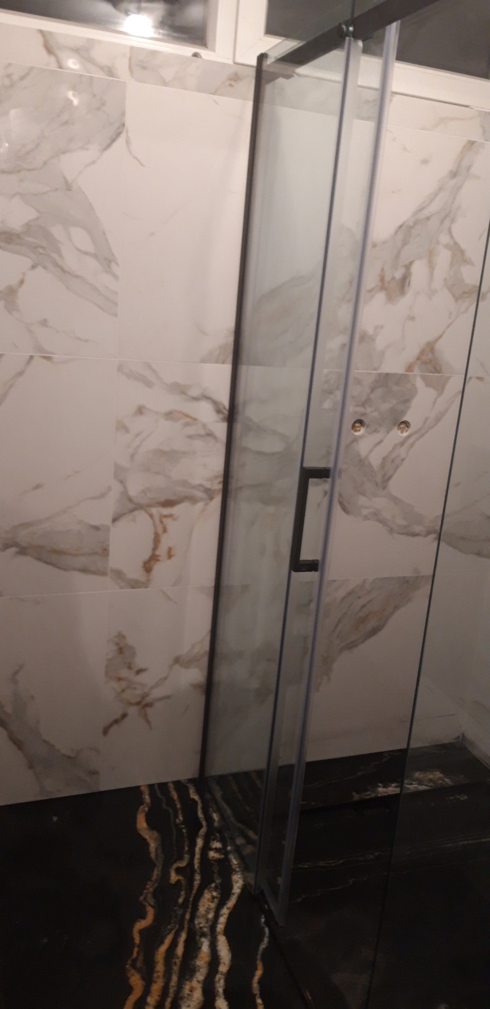 Szklana kabina prysznicowa z czarnym uchwytem na tle marmurowych płytek ściennych i podłogi z czarnego kamienia z złotymi żyłami.