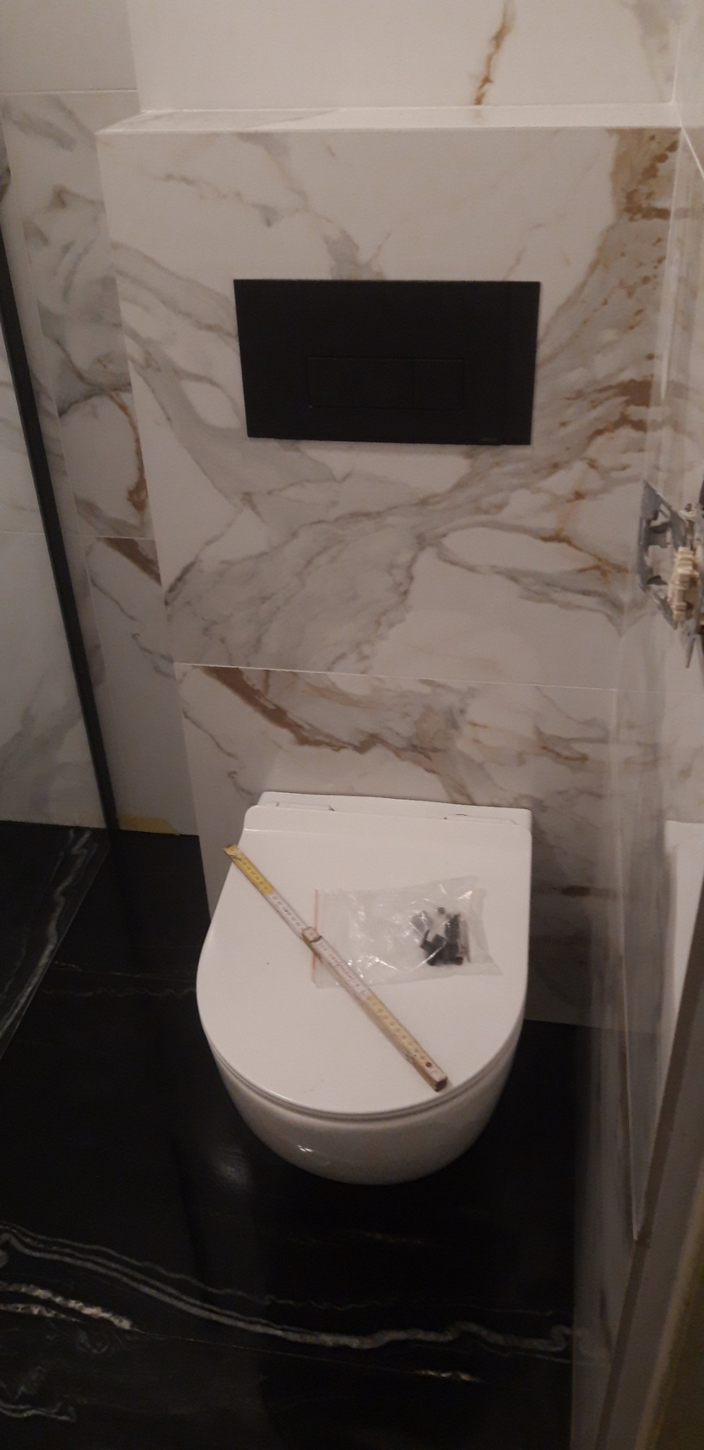 Wisząca miska WC z czarnym przyciskiem spłukującym na tle marmurowych płytek, na desce leżą akcesoria montażowe w folii i rozłożona miarka.