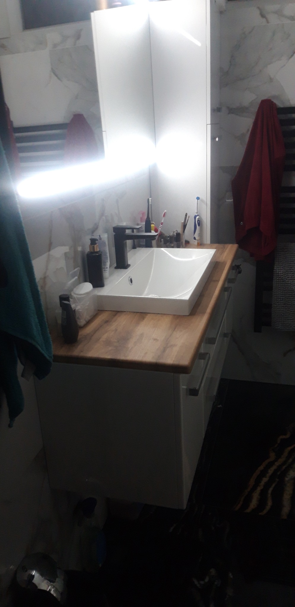 Biała umywalka nablatowa z czarną baterią na drewnopodobnym blacie szafki w łazience z marmurowymi płytkami, widoczne akcesoria łazienkowe i ręczniki.