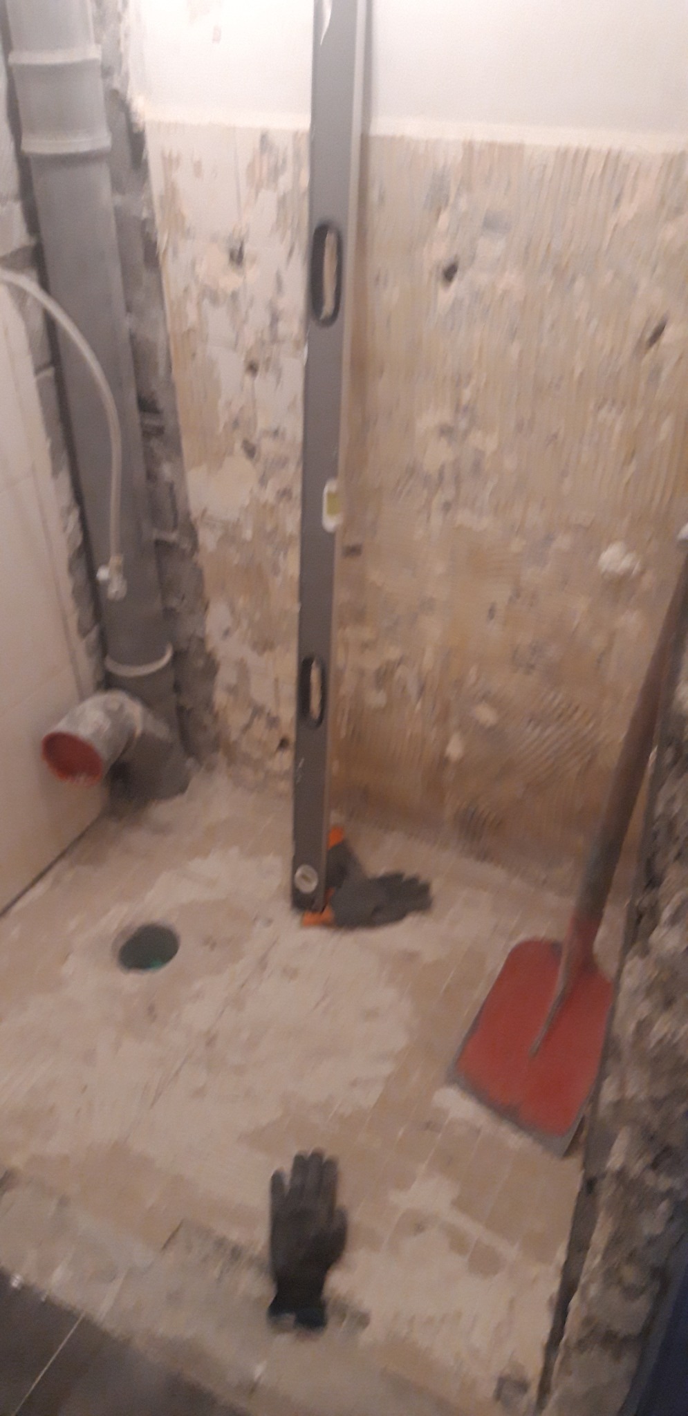 Przygotowanie podłoża w łazience: widoczne rury kanalizacyjne, poziomica oparta o ścianę, rękawice robocze i łopata, ściany częściowo przygotowane pod płytki.