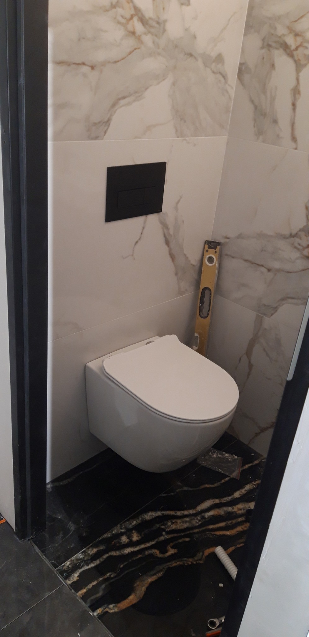 Wisząca toaleta z czarnym panelem sterującym na ścianie wyłożonej płytkami imitującymi marmur, poziomica oparta o ścianę, podłoga z czarnych płytek z jasnymi żyłami.