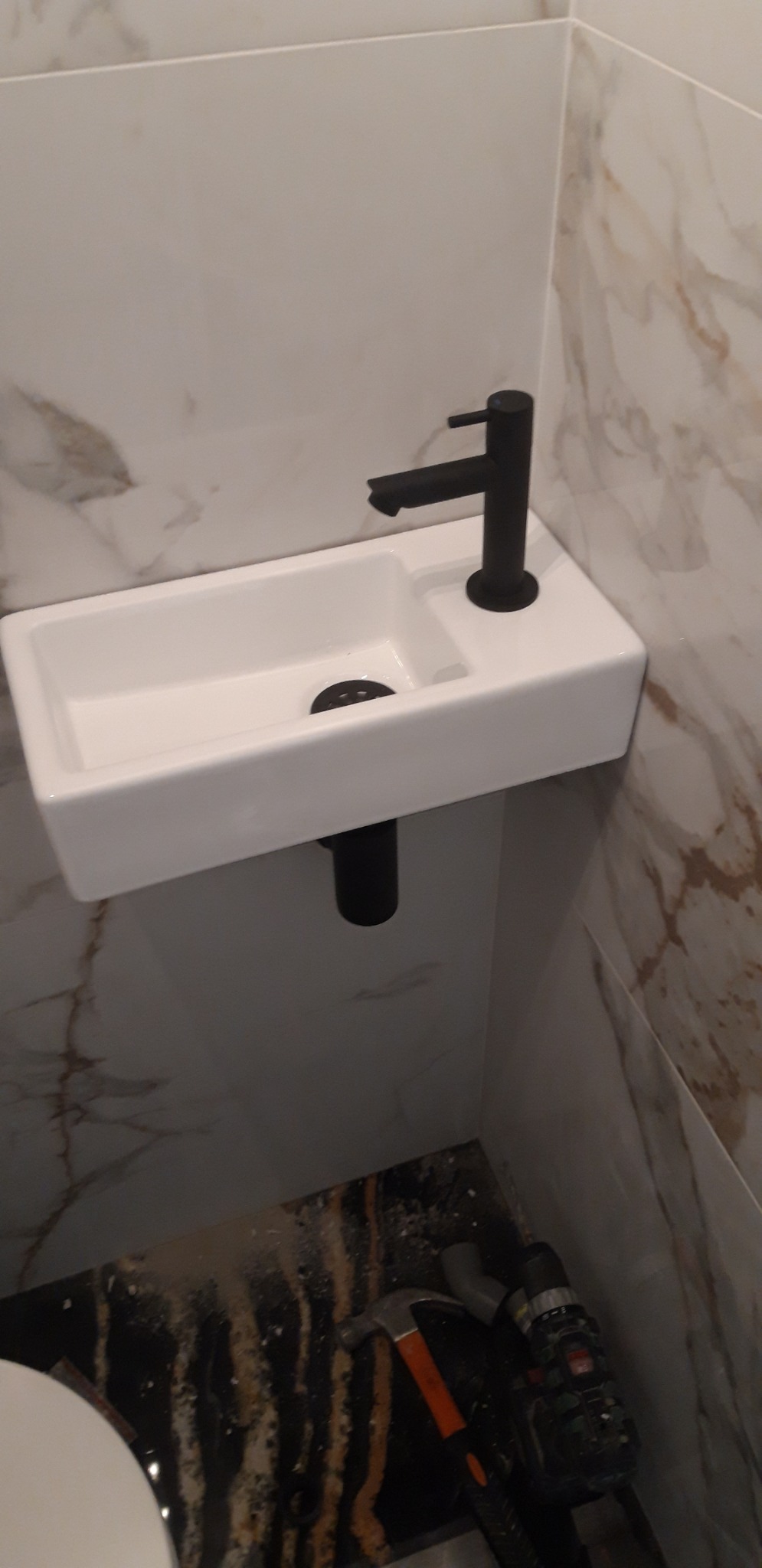 Mała, prostokątna umywalka z czarną baterią zamontowana na ścianie wyłożonej marmurowymi płytkami w Złotowie. Na podłodze widoczne narzędzia i fragment muszli klozetowej.