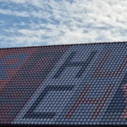 Ukończony dach z dachówki ceramicznej, na którym ułożono wzór z dachówek w dwóch kolorach: czerwonym i szarym, na tle błękitnego nieba z białymi obłokami.