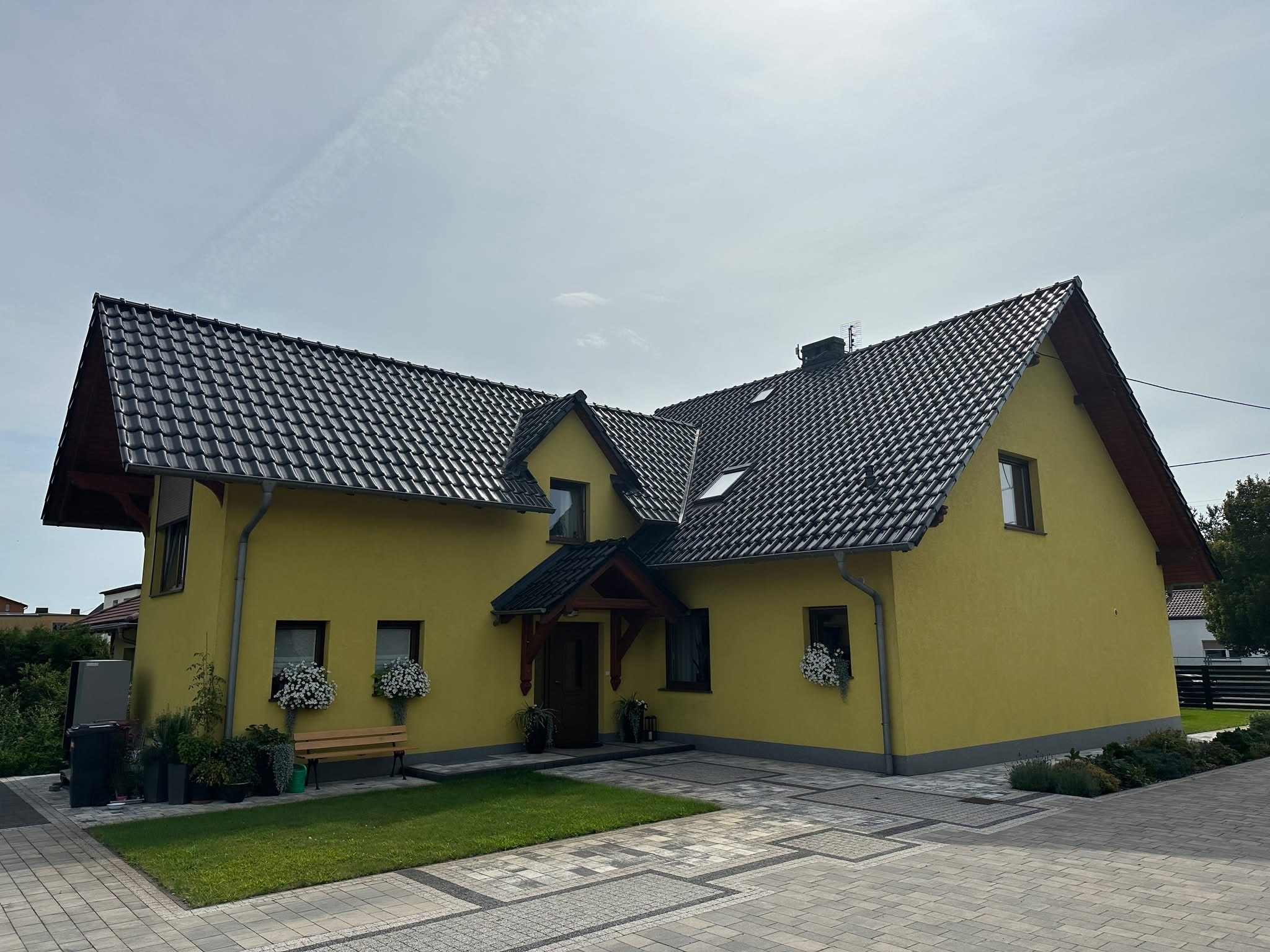 Żółty dom jednorodzinny z ciemną dachówką, widok z zewnątrz, kostka brukowa na podjeździe, kwiaty w oknach.