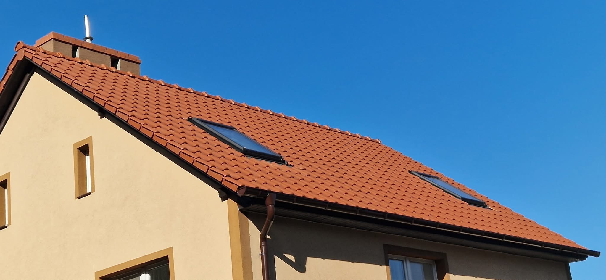 Czerwona dachówka ceramiczna z dwoma oknami dachowymi, brązowe rynny i komin na tle błękitnego nieba.