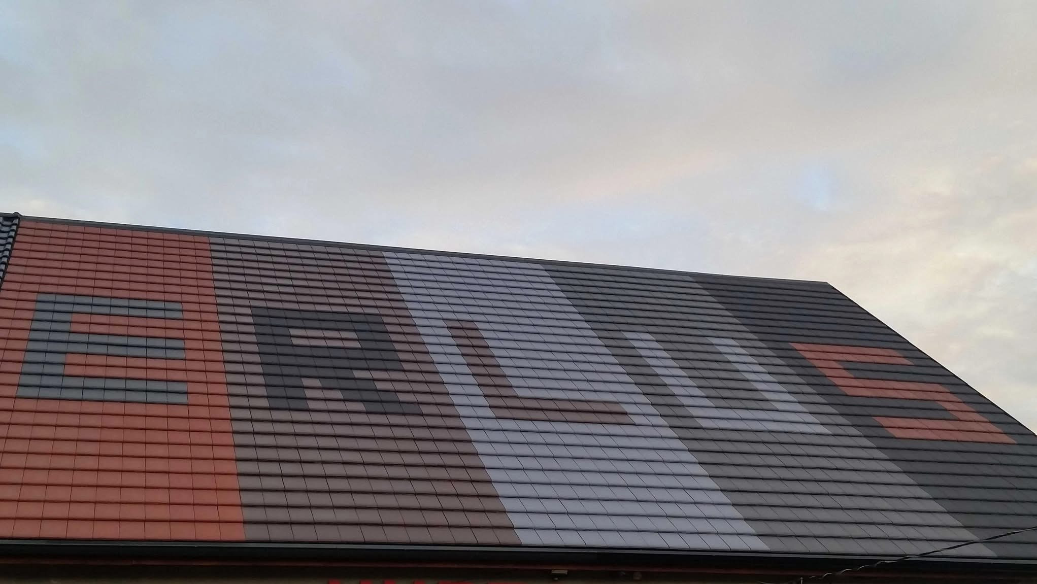 Dach pokryty dachówką ceramiczną, na której ułożono wzór w postaci dużych liter tworzących napis, widoczne z dołu pod kątem, pochmurne niebo w tle.