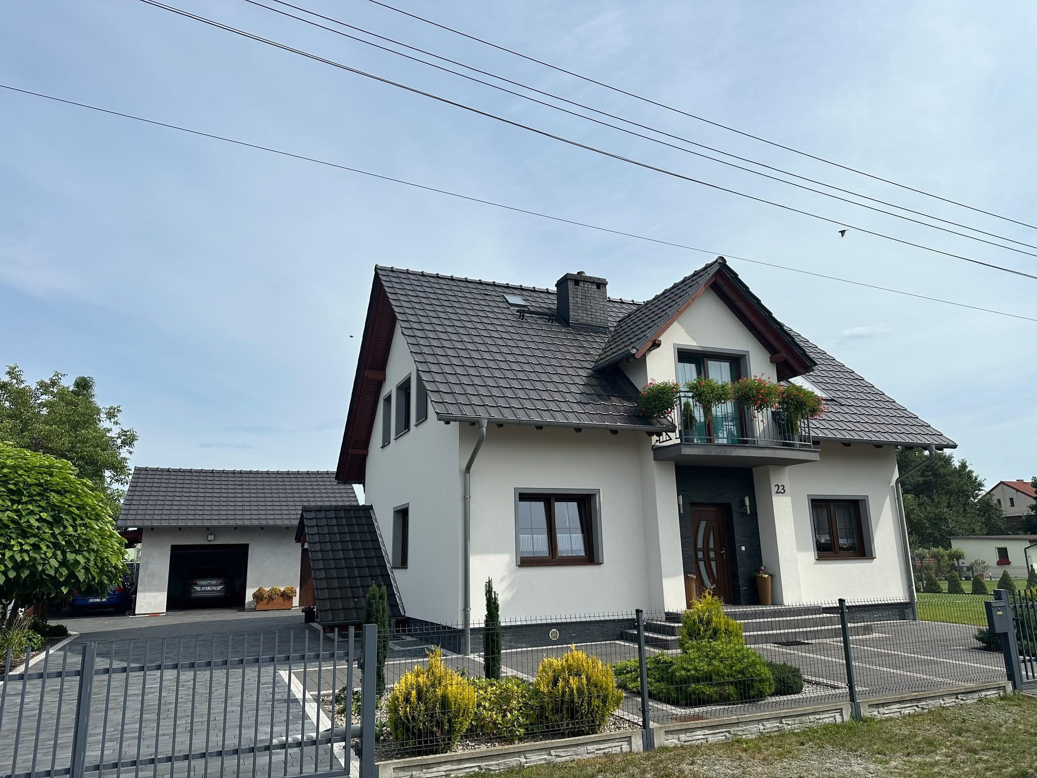 Nowoczesny dom jednorodzinny z szarą dachówką, balkonem z kwiatami i numerem 23, widoczny zza ogrodzenia.