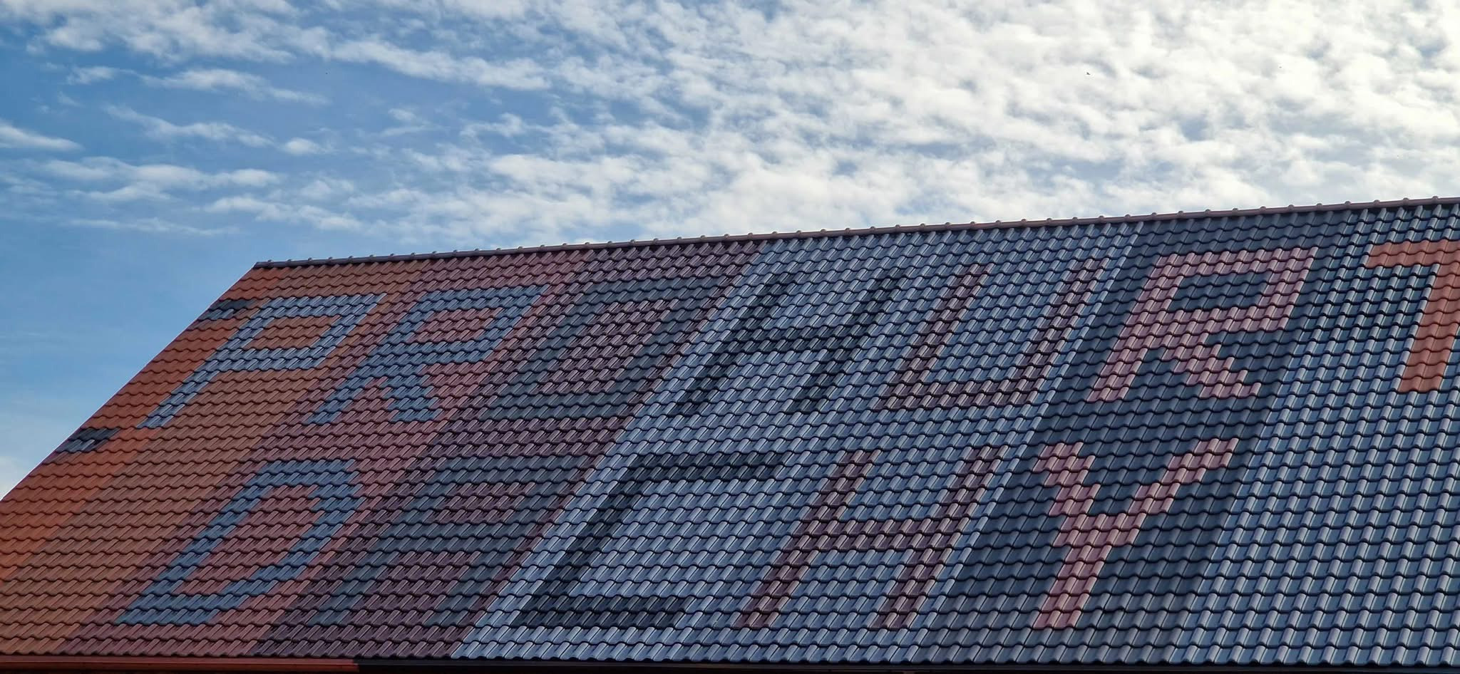 Ukończony dach z dachówki ceramicznej, na którym ułożono wzór z dachówek w dwóch kolorach: czerwonym i szarym, na tle błękitnego nieba z białymi obłokami.