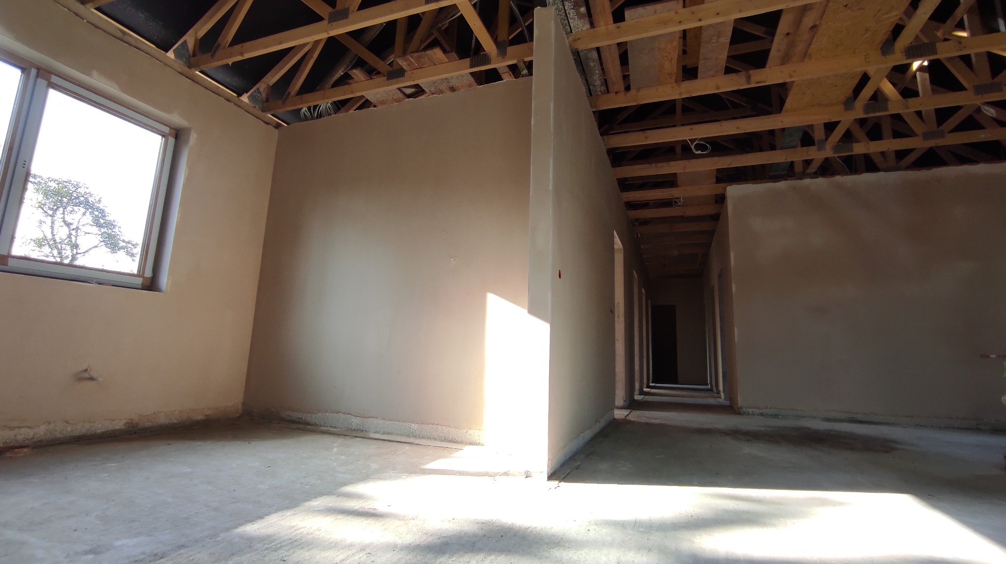 Surowe wnętrze w trakcie prac wykończeniowych; ściany pokryte jasnym, beżowym tynkiem, widoczna drewniana konstrukcja dachu i surowy beton na podłodze, z oknem po lewej stronie kadru.