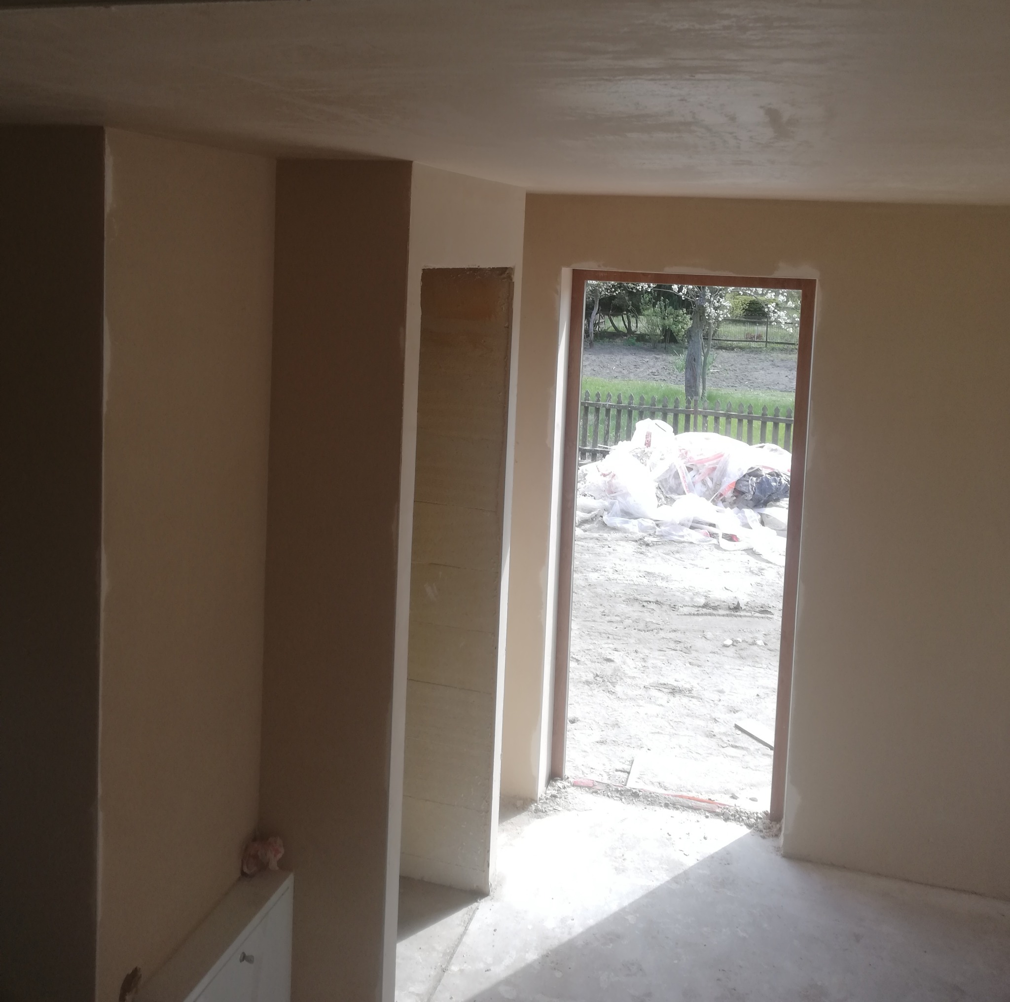 Wnętrze w trakcie prac wykończeniowych; ściany pokryte jasnym tynkiem, widoczna surowa powierzchnia wnęki, na zewnątrz okna stos materiałów budowlanych.