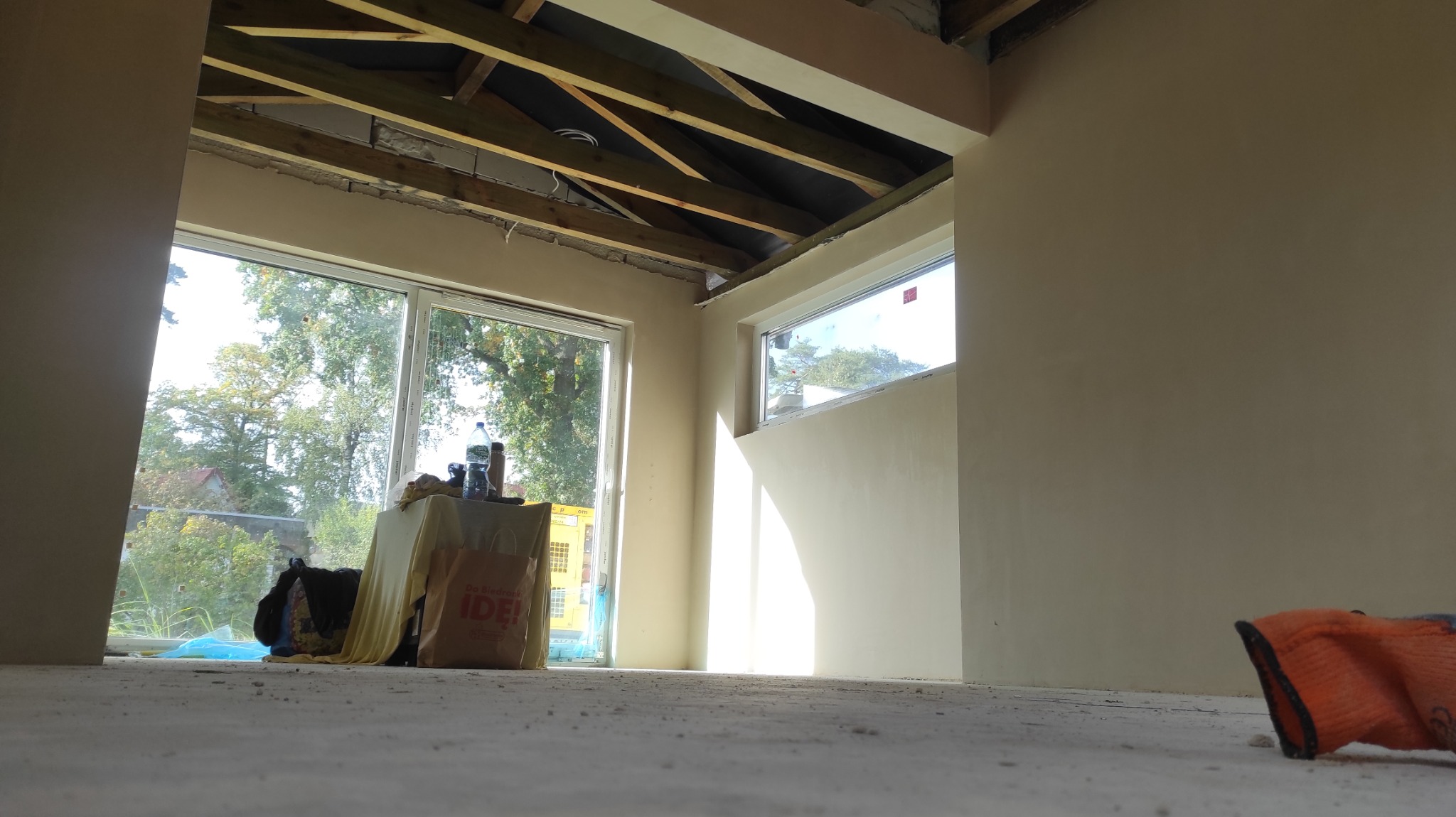 Wnętrze w trakcie prac wykończeniowych, świeżo nałożone jasne tynki na ścianach, widoczne okna z widokiem na zieleń, surowy drewniany strop i narzędzia pracy.