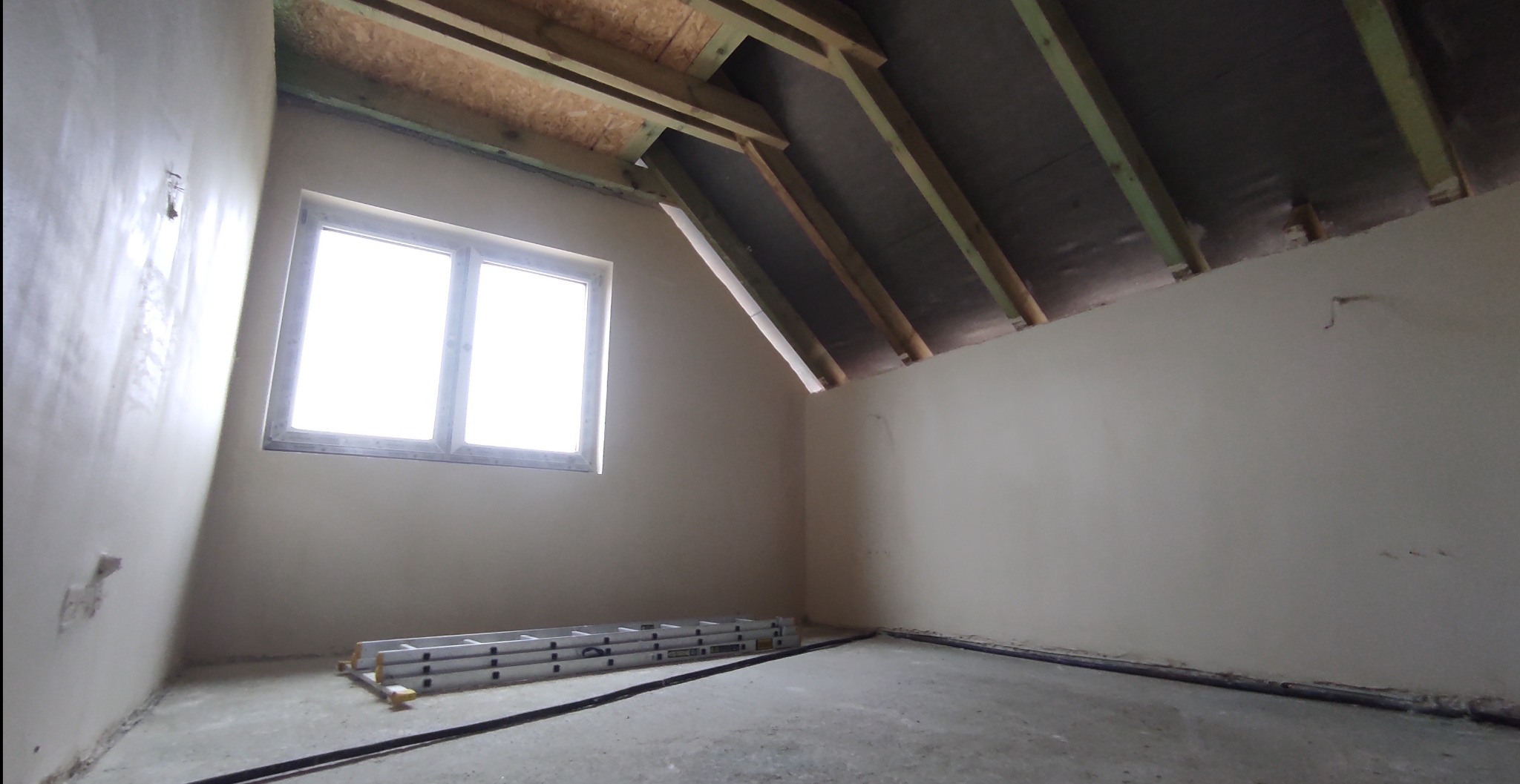 Poddasze ze skośnym dachem, ściany pokryte świeżo nałożoną warstwą wykończeniową, aluminiowa drabina oparta o ścianę pod oknem.