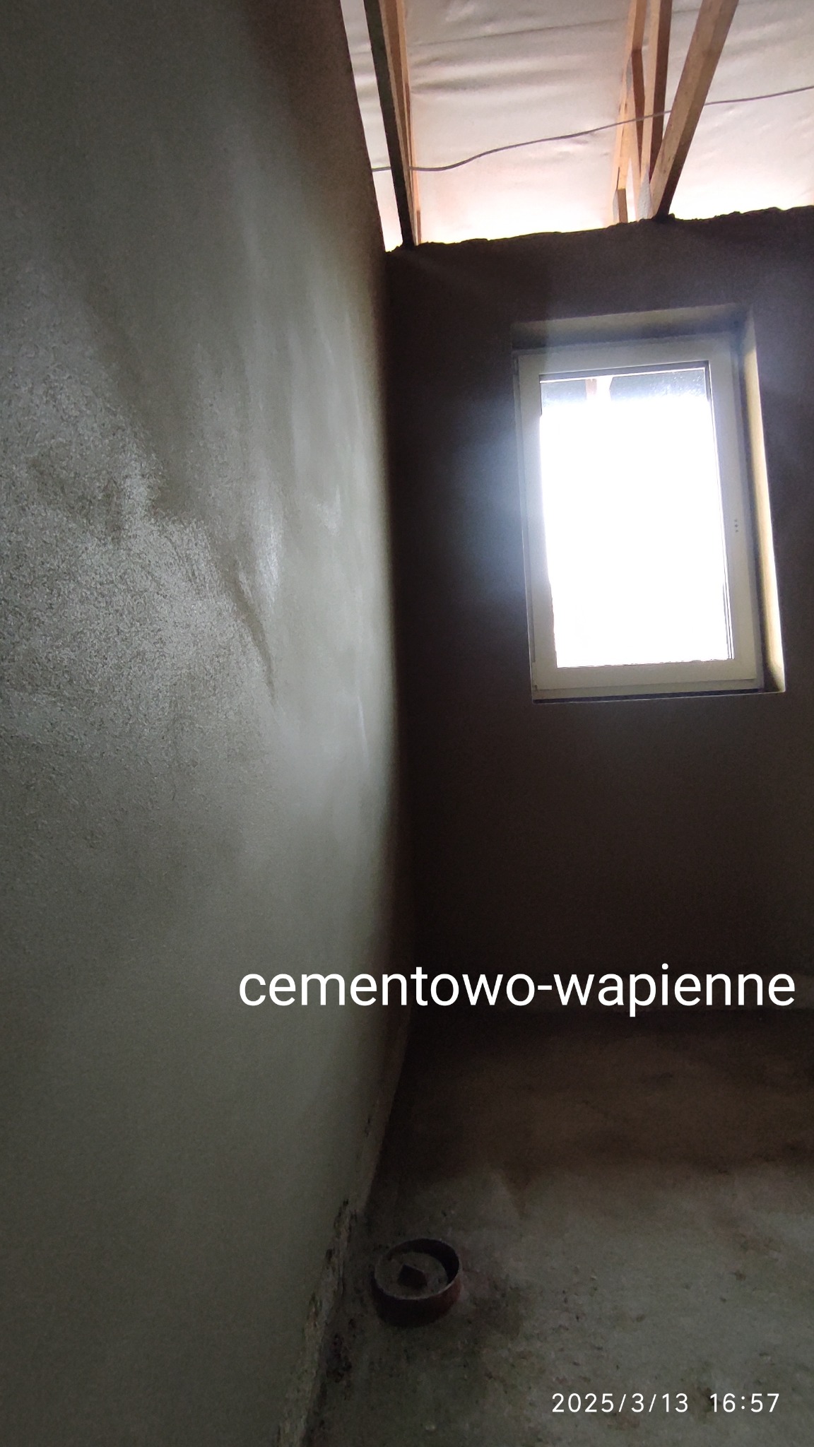 Świeżo nałożona, wilgotna warstwa cementowo-wapienna na ścianie w surowym wnętrzu, widoczne drewniane elementy konstrukcji dachu i okno w tle.