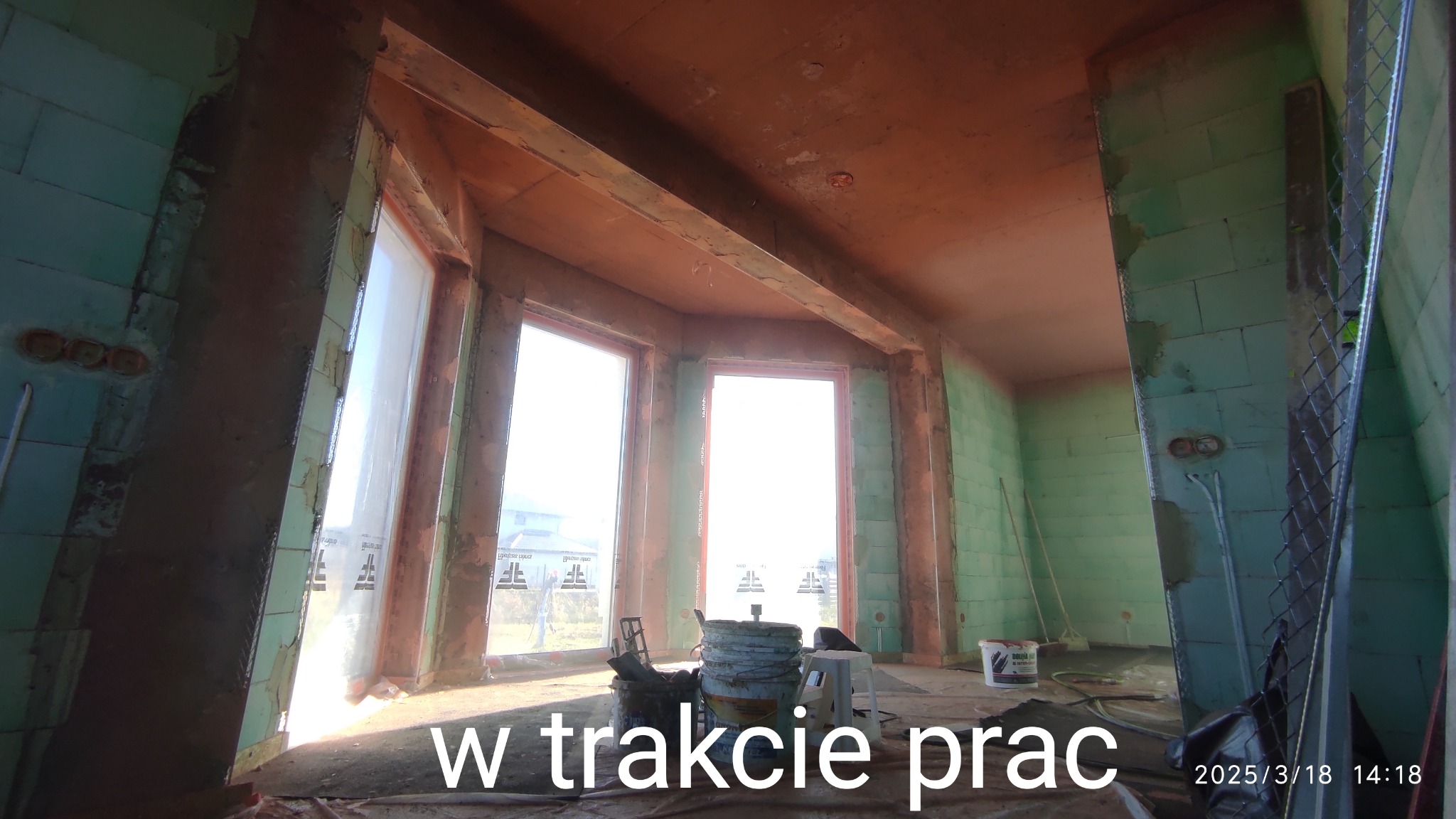 Wnętrze w trakcie prac wykończeniowych, ściany z zielonych bloczków i sufit pokryte szarobrązową warstwą, widoczne wiadra i narzędzia na podłodze, duże okna z widokiem na zewnątrz.