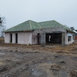 Piotr Faszyński - Dom w trakcie budowy z częściowo położonym zielonym pokryciem dachowym na łacie, widoczne rusztowania i błotniste podłoże.