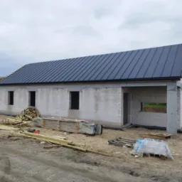 Surowy budynek w trakcie budowy z nowym, czarnym dachem z blachy na rąbek stojący, widoczne materiały budowlane, teren przygotowany pod dalsze prace.
