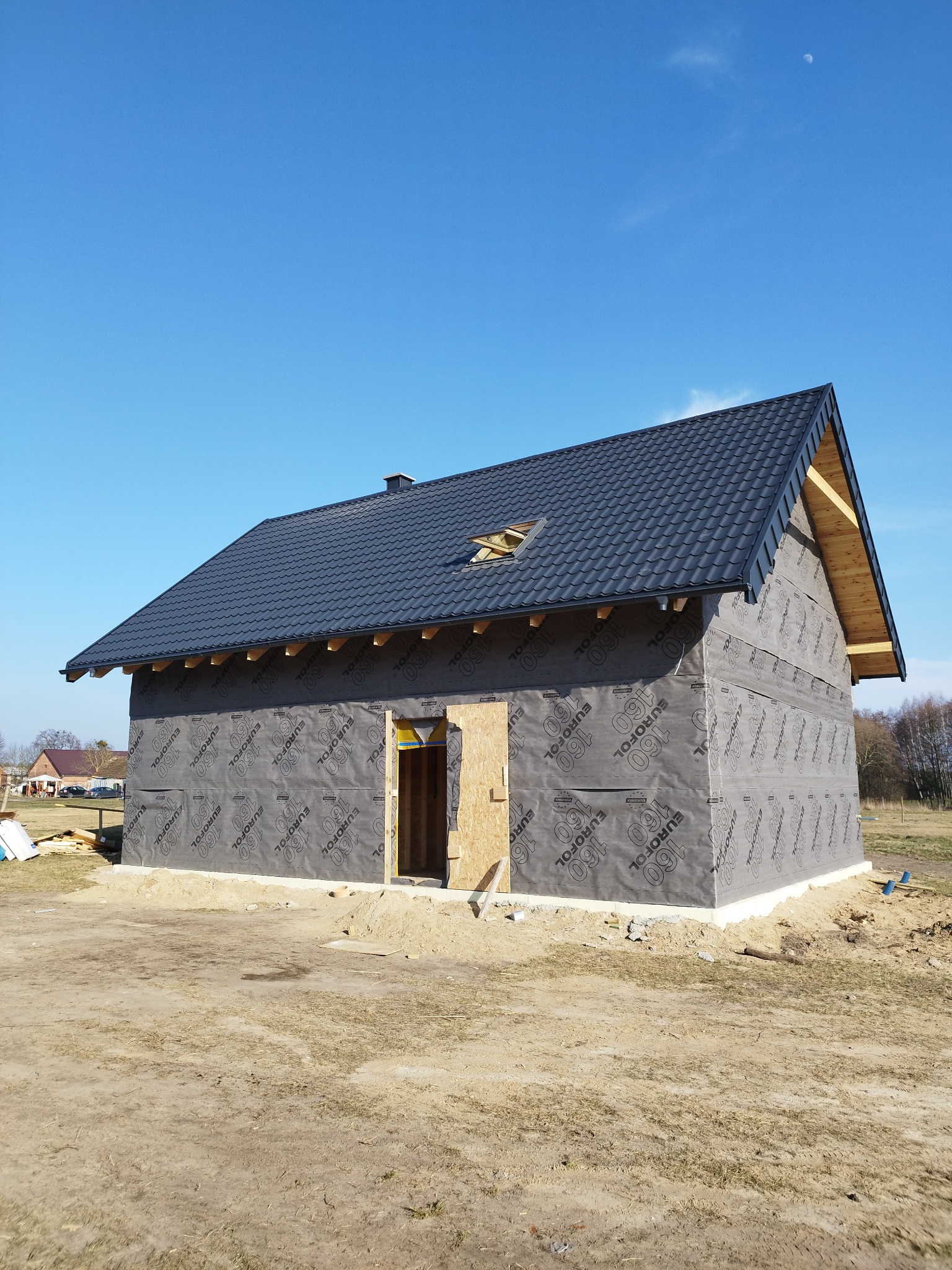 Nowy dom w trakcie budowy z czarnym dachem z dachówki, zabezpieczony folią izolacyjną z nadrukiem, widoczne przygotowane wejście bez drzwi i niebo z księżycem w tle.