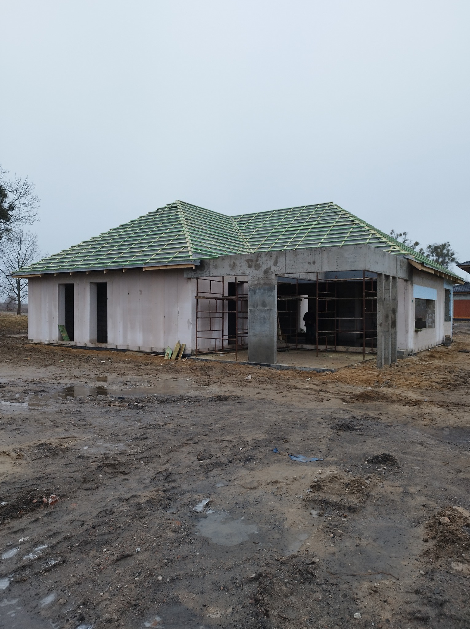 Dom w trakcie budowy z częściowo położonym zielonym pokryciem dachowym na łacie, widoczne rusztowania i błotniste podłoże.