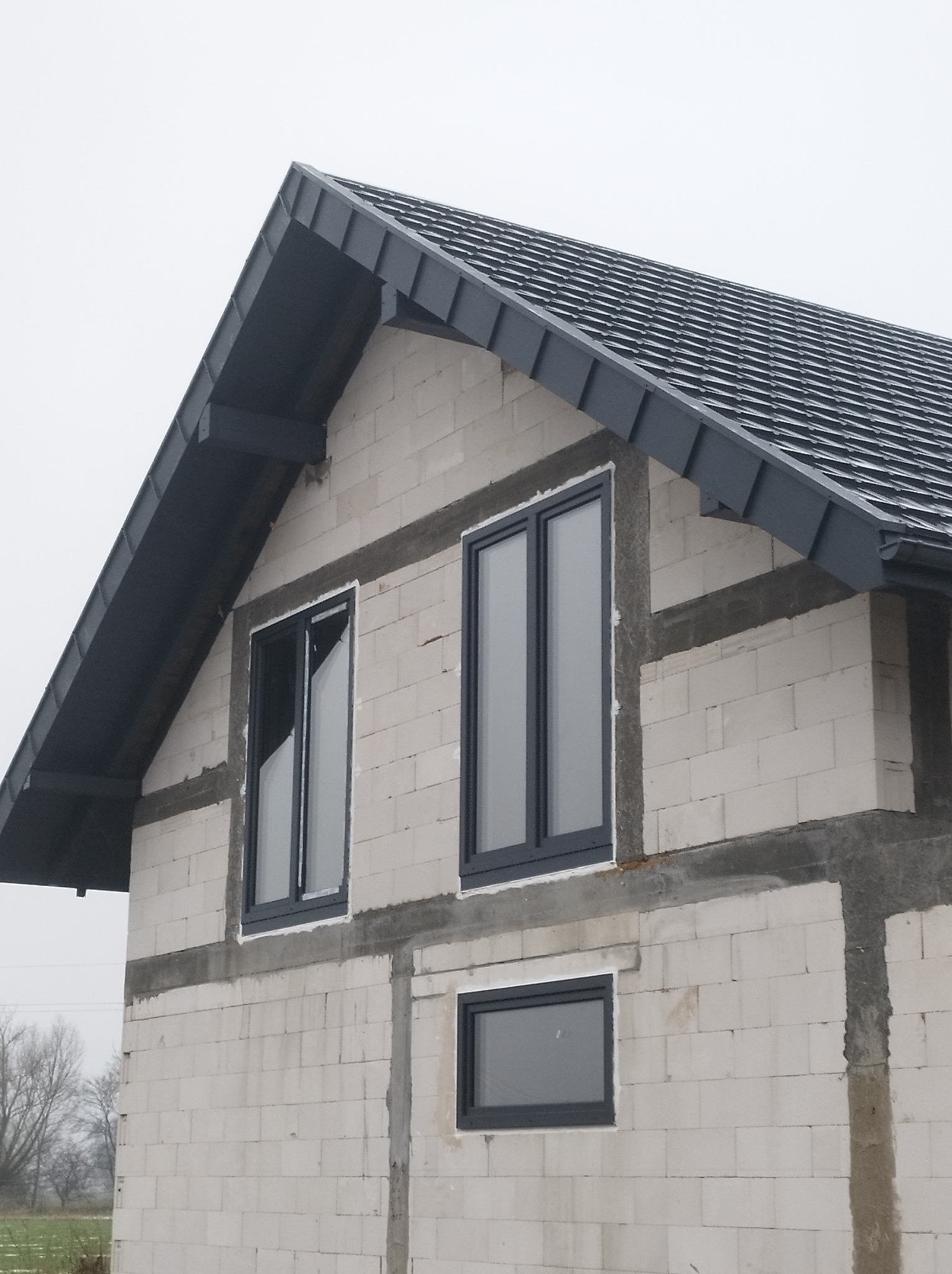 Surowy stan budynku z białego pustaka z zamontowaną, ciemnoszarą blachodachówką i oknami w ciemnych ramach, widok z dołu na tle pochmurnego nieba.