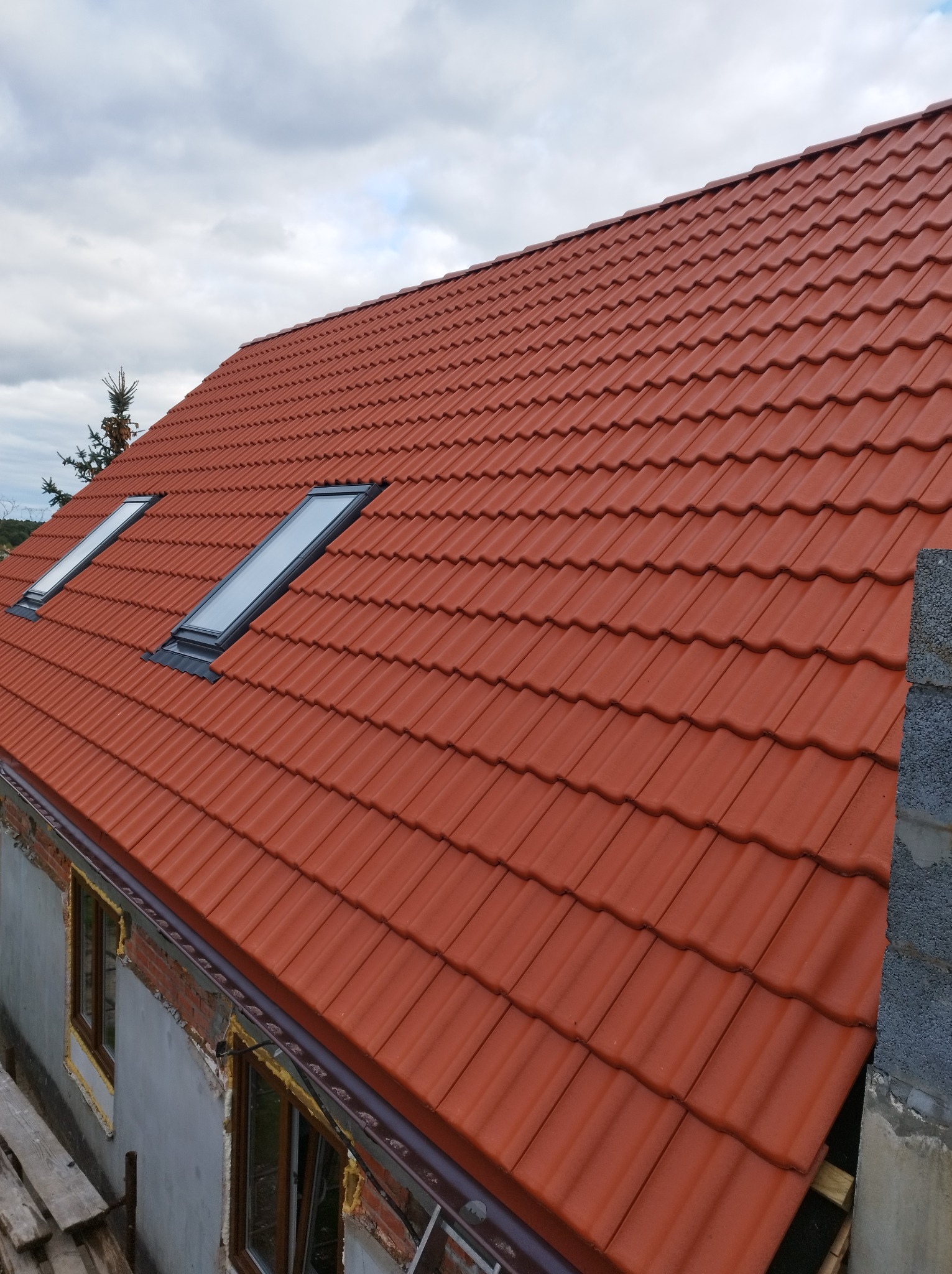 Czerwony dach z dachówki ceramicznej z dwoma oknami dachowymi, widoczne niedokończone obrzeża okien i komina, fragment ściany budynku i pochmurne niebo.