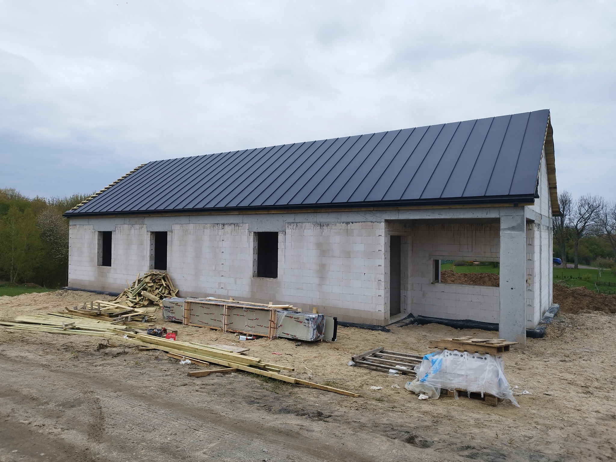 Surowy budynek w trakcie budowy z nowym, czarnym dachem z blachy na rąbek stojący, widoczne materiały budowlane, teren przygotowany pod dalsze prace.