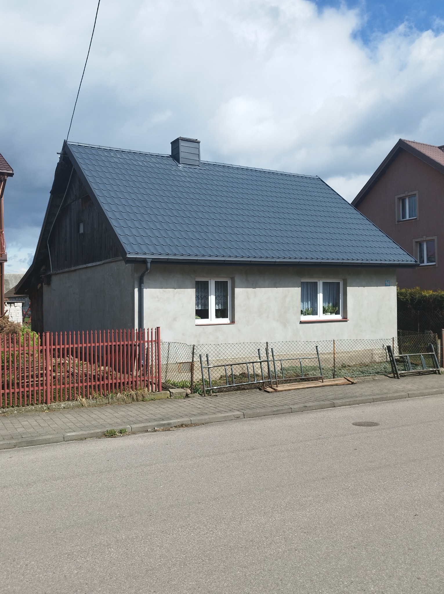 Niski dom jednorodzinny z szarą blachodachówką i szarym kominem, widoczny z ulicy, z drabinkami opartymi o ogrodzenie.