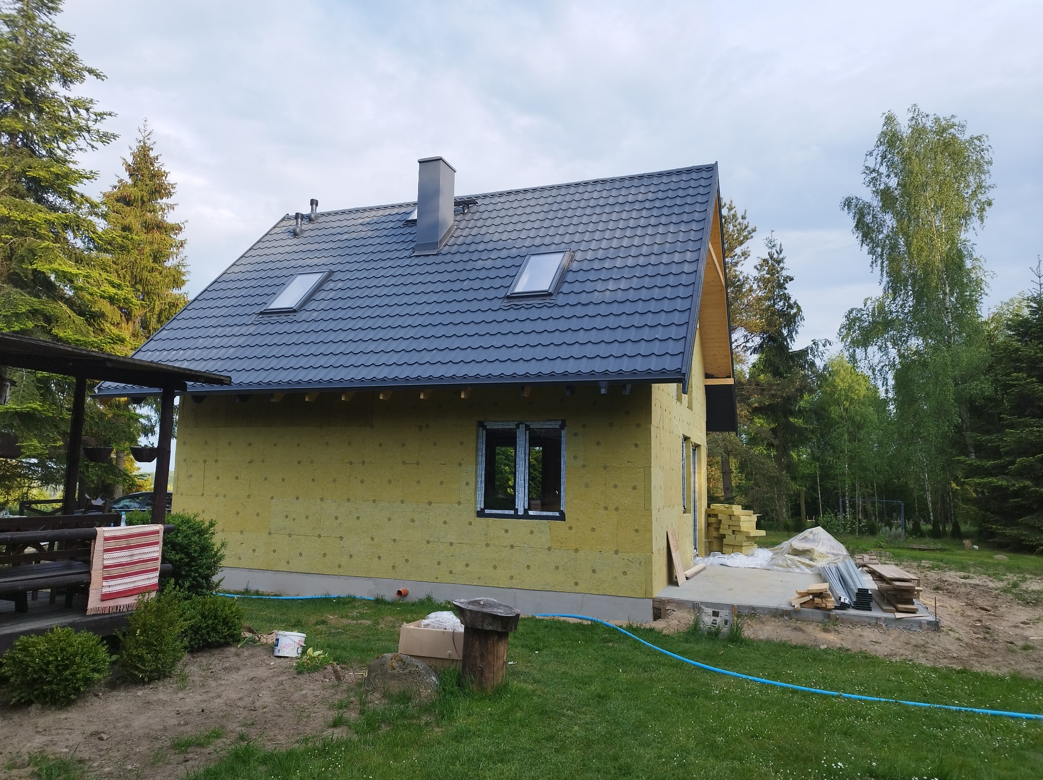 Dom w trakcie ocieplania, z nowym, grafitowym dachem z oknami dachowymi i kominem, ściany pokryte żółtą wełną mineralną, widoczne materiały budowlane na zewnątrz.