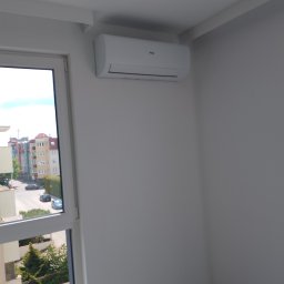 KLIMACIK ADRIAN WOJCIECHOWSKI - Biały klimatyzator Haier zamontowany na ścianie w jasnym pokoju, widok z okna na budynki mieszkalne i zieleń. Widoczny fragment gniazdka elektrycznego.