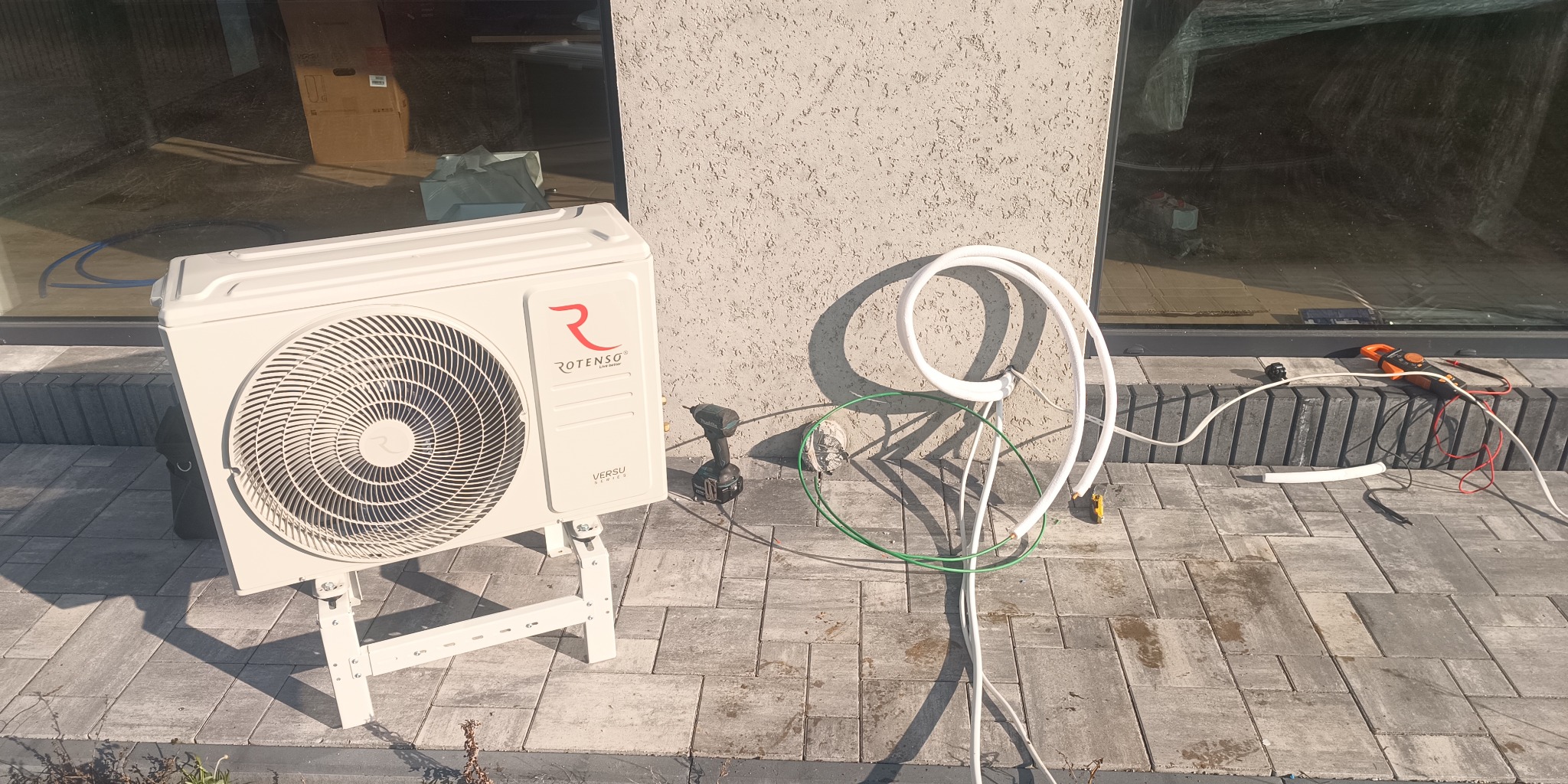 Zewnętrzna jednostka klimatyzacji Rotenso Versu na białym stelażu, obok wiertarka akumulatorowa Makita i przewody instalacyjne na brukowanym tarasie, ściana budynku z fakturą baranka w tle.