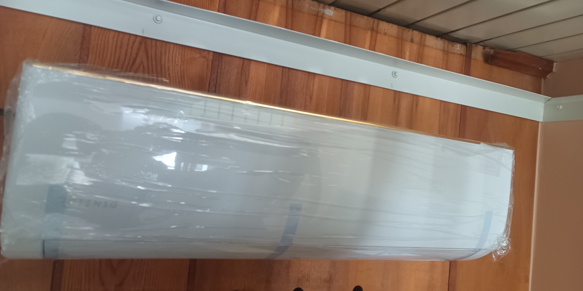 Nowy klimatyzator marki 'TENSO' jeszcze w folii ochronnej, zamontowany na drewnianej ścianie pod sufitem z listwą maskującą.
