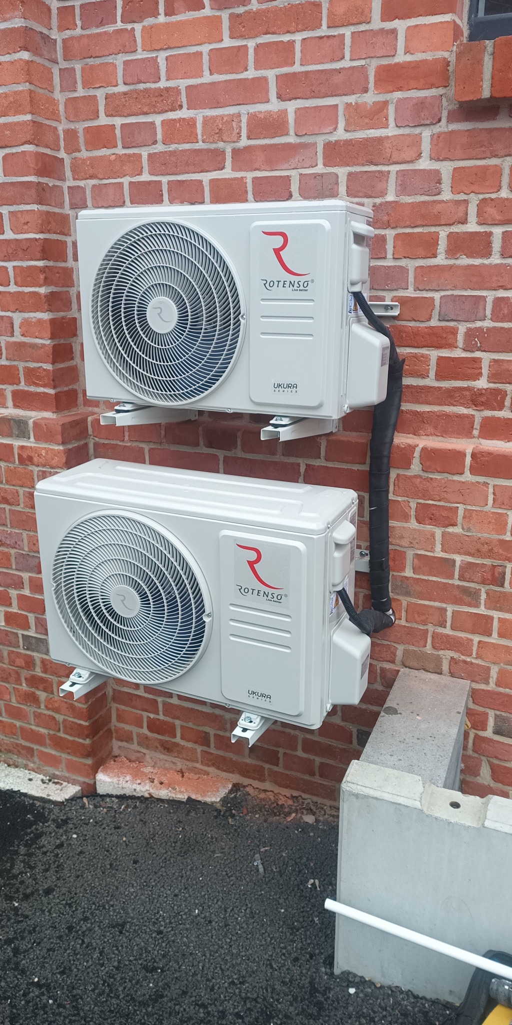 Dwa białe klimatyzatory Rotenso Ukura zamontowane jeden nad drugim na czerwonej ceglanej ścianie zewnętrznej budynku.