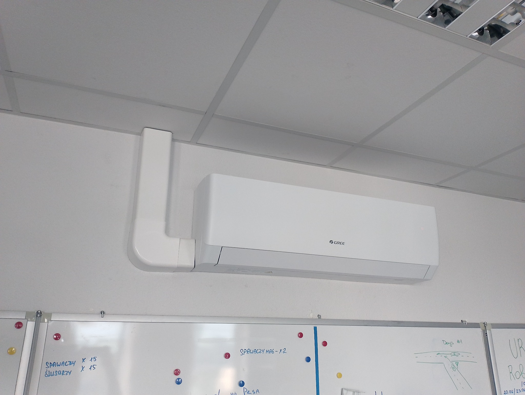 Biała klimatyzacja Gree zamontowana na ścianie w pomieszczeniu biurowym z widoczną tablicą suchościeralną z notatkami. Widoczne rury instalacyjne. Oświetlenie sufitowe.
