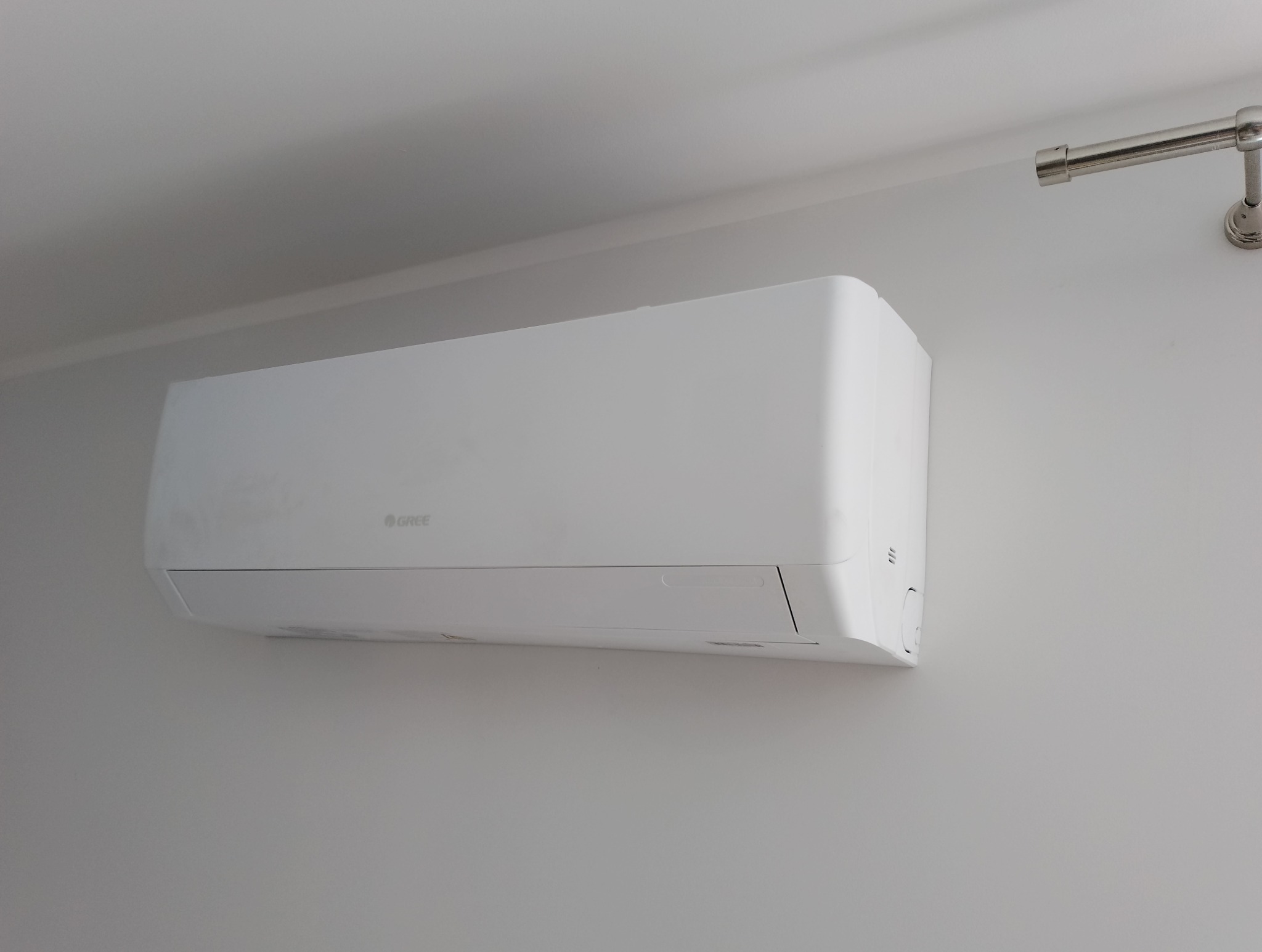 Biały klimatyzator marki Gree zamontowany na jasnej ścianie, widoczny fragment sufitu i karnisza. Minimalistyczny design.