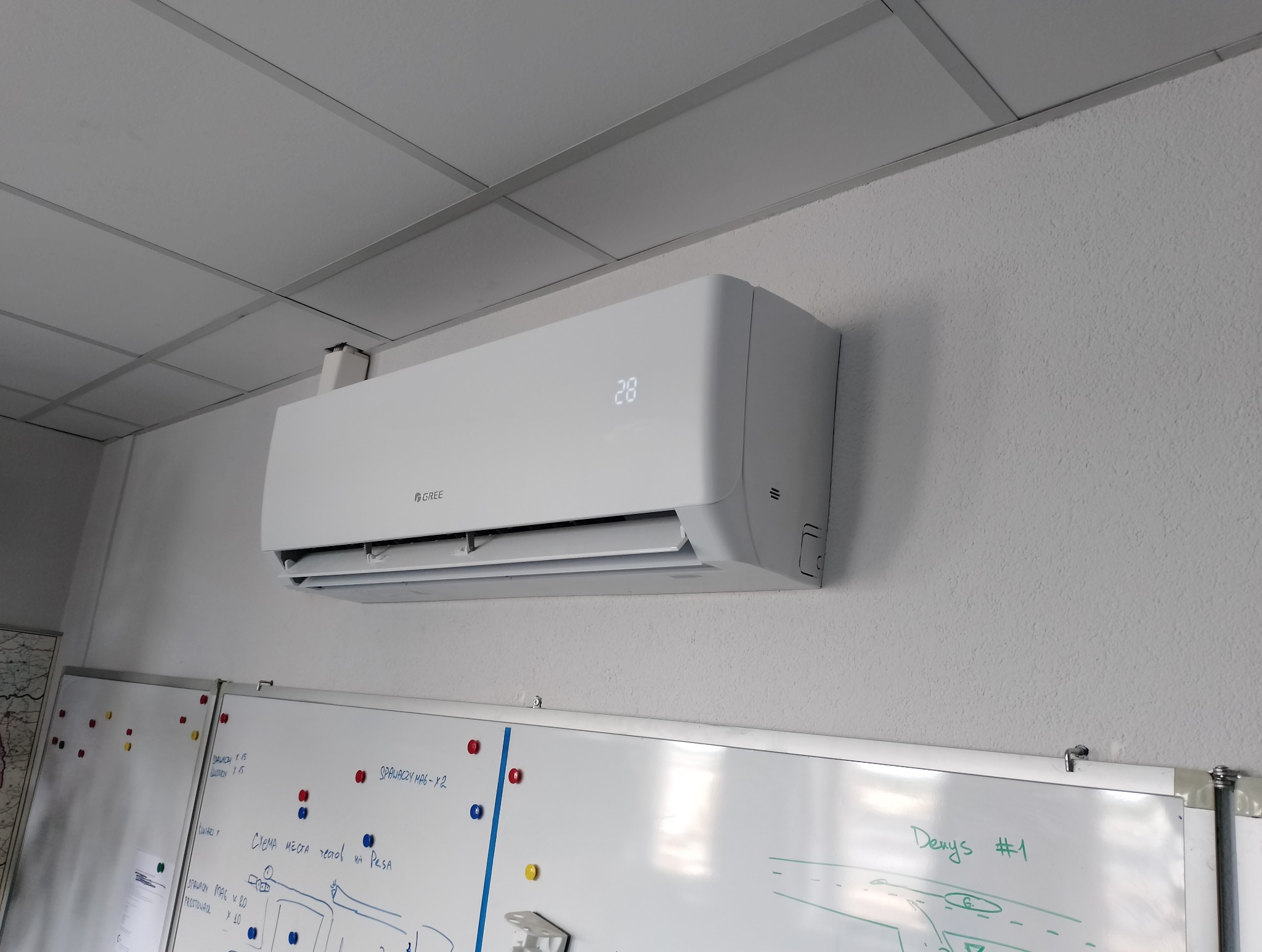 Biała klimatyzacja Gree zamontowana na ścianie biura, z tablicami suchościeralnymi poniżej. Widoczny wyświetlacz temperatury. Oświetlenie sufitowe rastrowe.