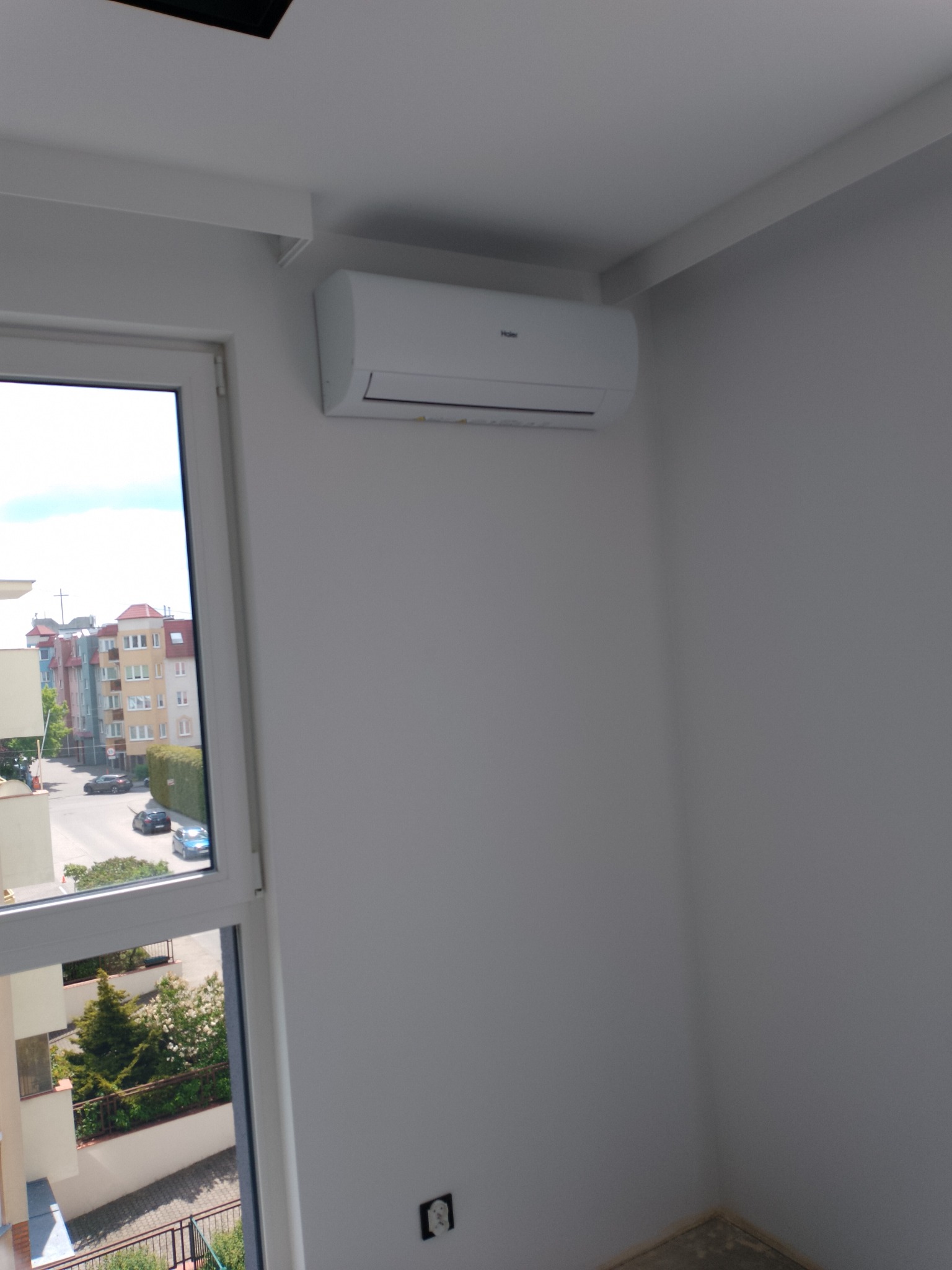 Biały klimatyzator Haier zamontowany na ścianie w jasnym pokoju, widok z okna na budynki mieszkalne i zieleń. Widoczny fragment gniazdka elektrycznego.