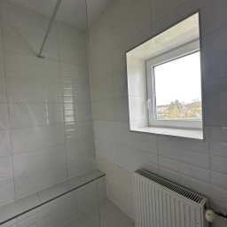 Usługi Remontowo-Budowlane DANIEL SOKOŁOWSKI-RYCHLIŃSKI - Wykończona łazienka w jasnych odcieniach szarości z kabiną prysznicową typu walk-in, grzejnikiem żeberkowym pod oknem i płytkami imitującymi cegłę.