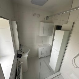 Usługi Remontowo-Budowlane DANIEL SOKOŁOWSKI-RYCHLIŃSKI - Jasna łazienka z białymi płytkami, umywalką na postumencie, grzejnikiem żeberkowym, szafkami wiszącymi i stojącymi oraz kabiną prysznicową ze szklaną ścianką.