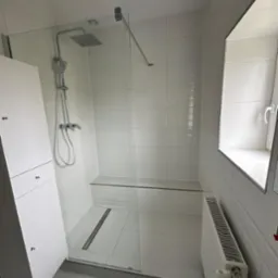 Nowoczesna kabina prysznicowa walk-in z odpływem liniowym, wykończona białymi płytkami, z siedziskiem i grzejnikiem pod oknem.