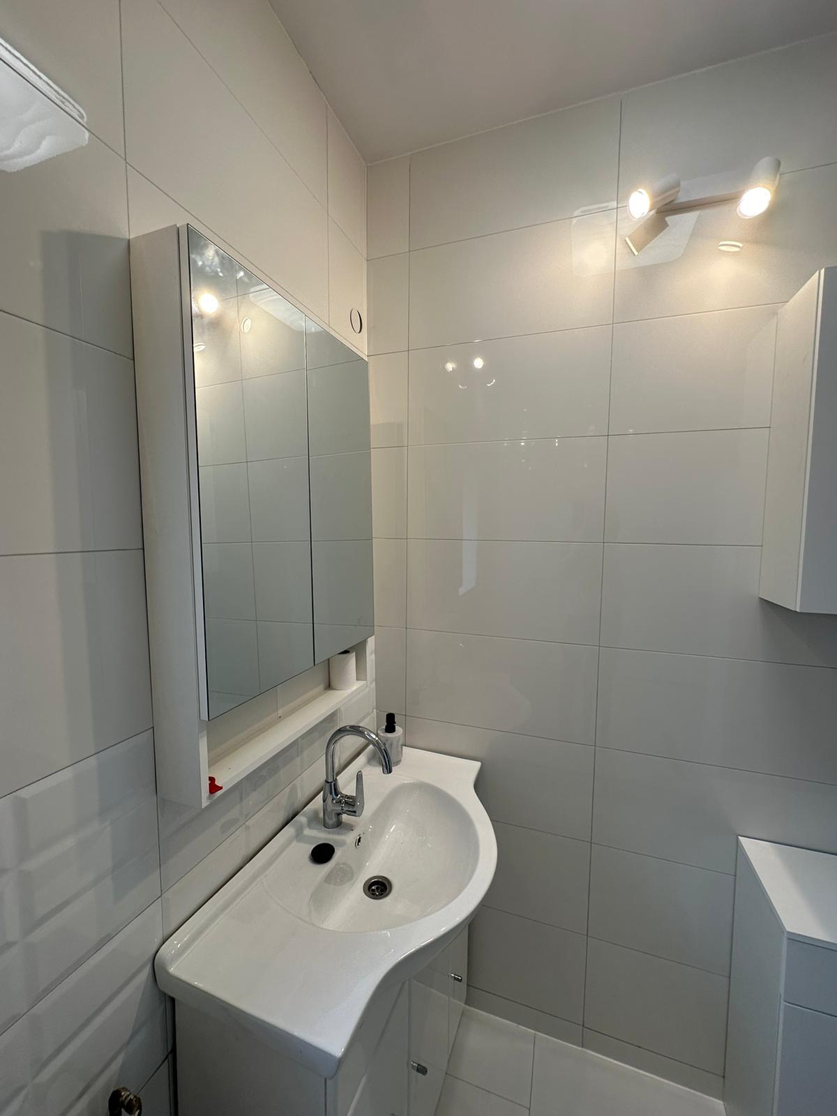 Biała umywalka z szafką i lustrem w nowoczesnej, wykafelkowanej łazience z oświetleniem punktowym.