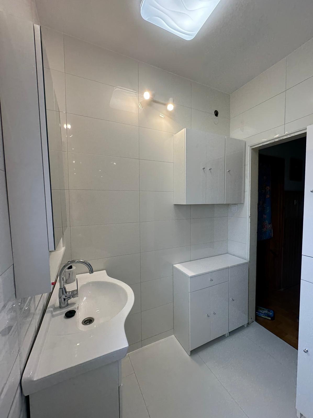 Jasna łazienka po remoncie: białe płytki, umywalka z szafką, wisząca szafka z trzema drzwiczkami i szafka pod umywalkę z szufladą. Widoczne także oświetlenie sufitowe i lustro.