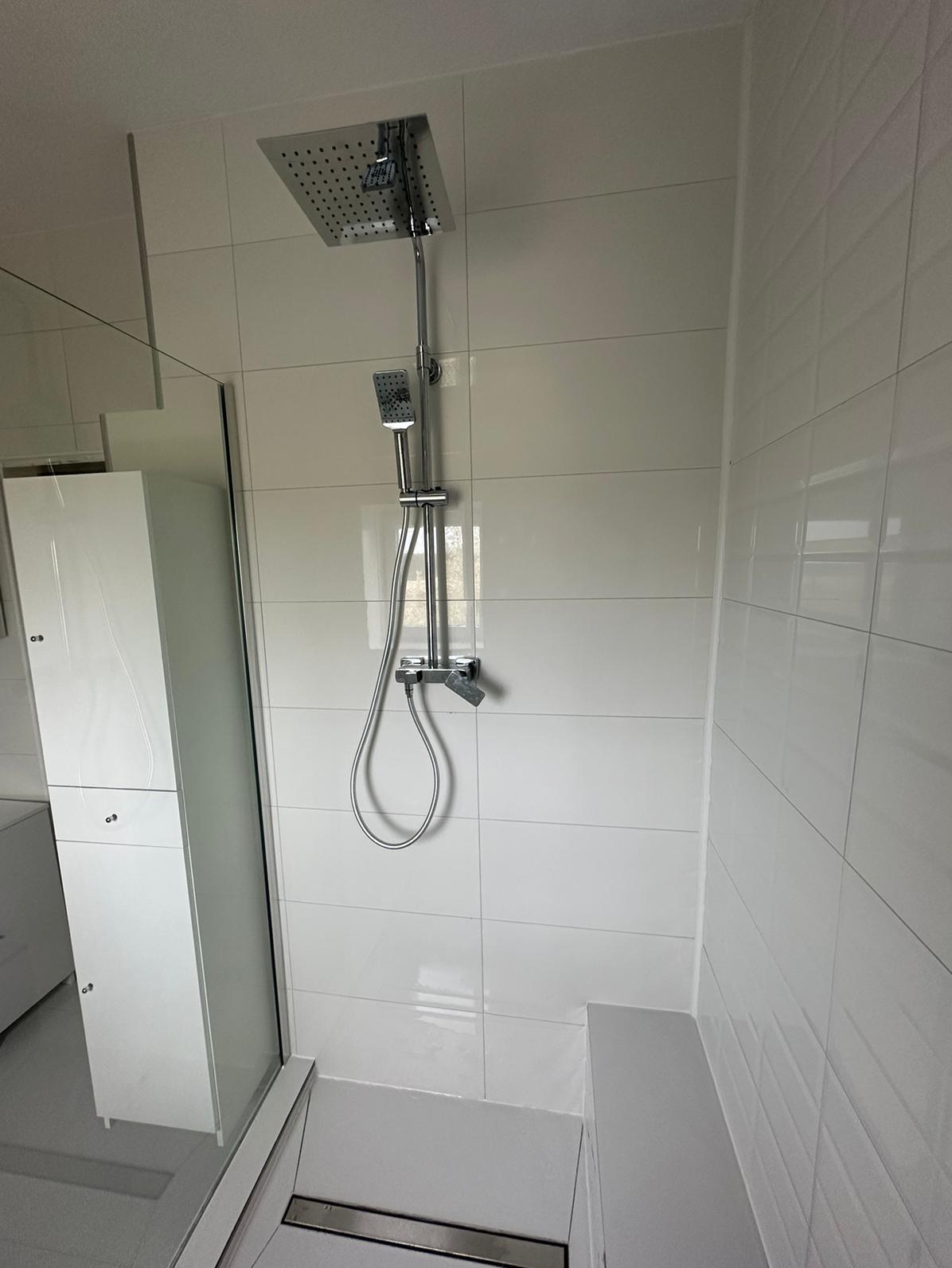 Nowoczesna kabina prysznicowa z deszczownicą i słuchawką, wykończona białymi, błyszczącymi płytkami w układzie prostokątnym, z widocznym odpływem liniowym i szafką łazienkową w tle.