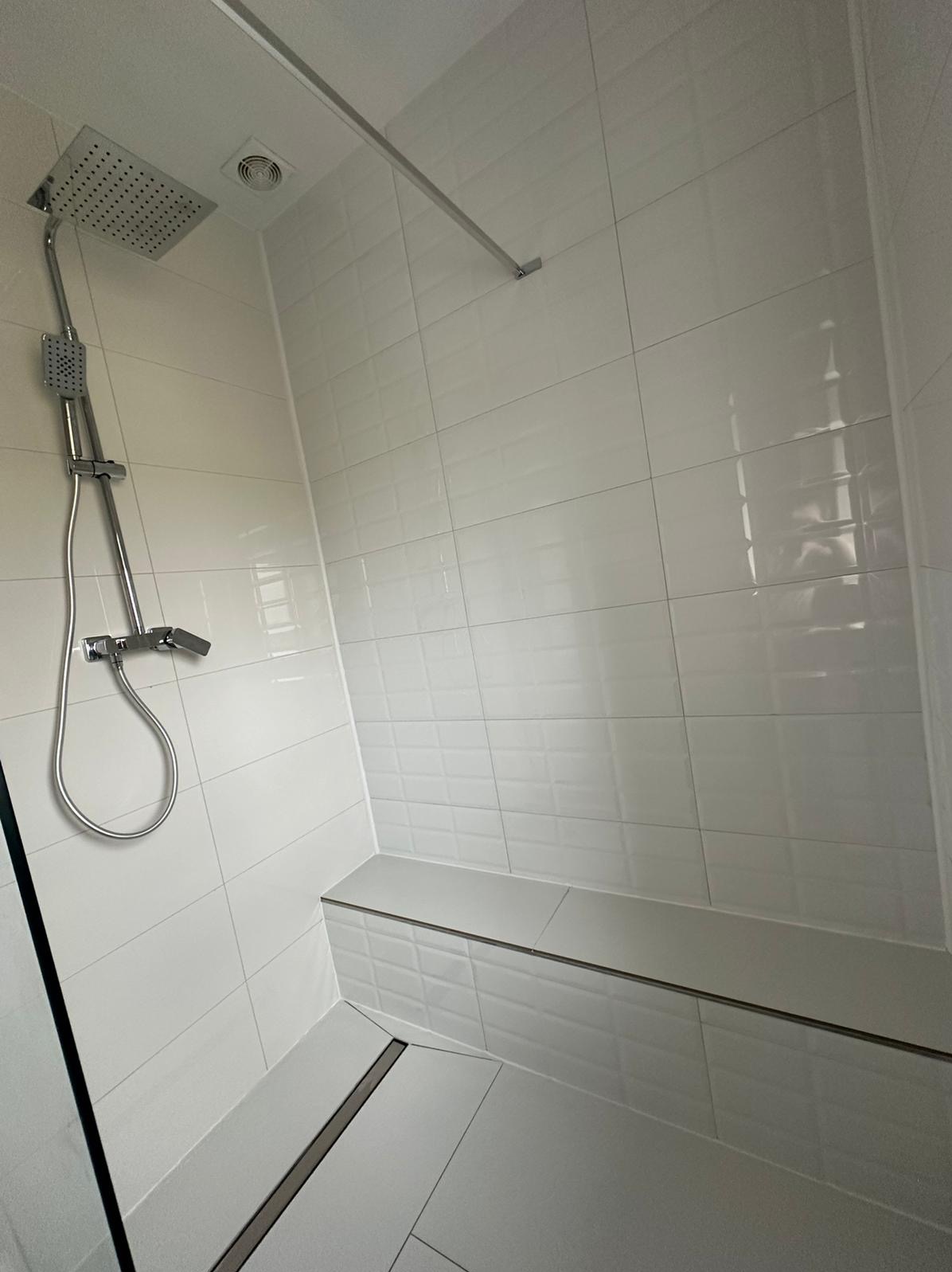 Nowoczesna kabina prysznicowa z białą, cegiełkową glazurą, deszczownicą i odpływem liniowym w podłodze, widoczne siedzisko wyłożone tymi samymi płytkami.