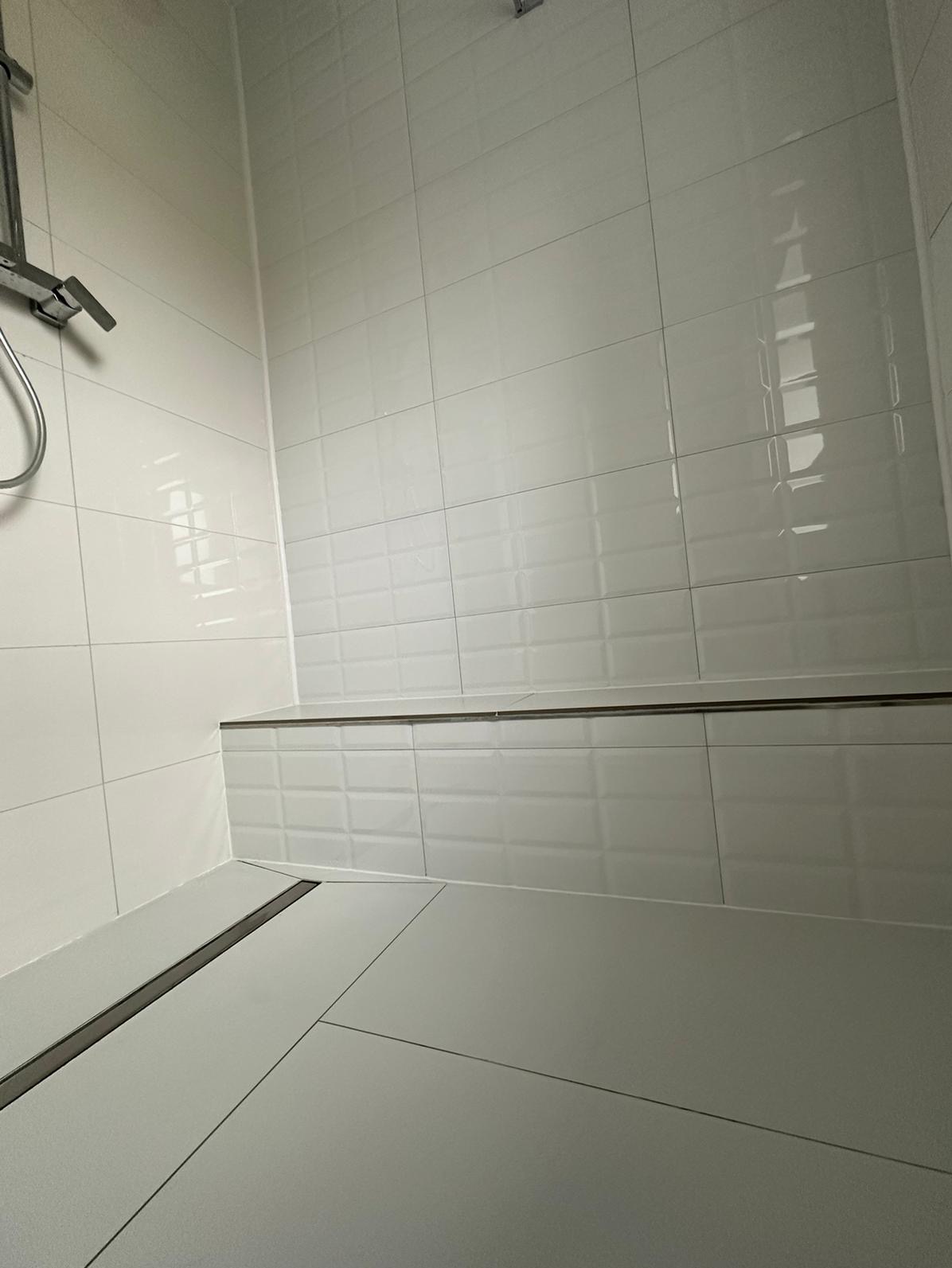 Nowoczesna kabina prysznicowa z białymi, błyszczącymi płytkami typu metro, szara podłoga z odpływem liniowym i chromowana armatura.