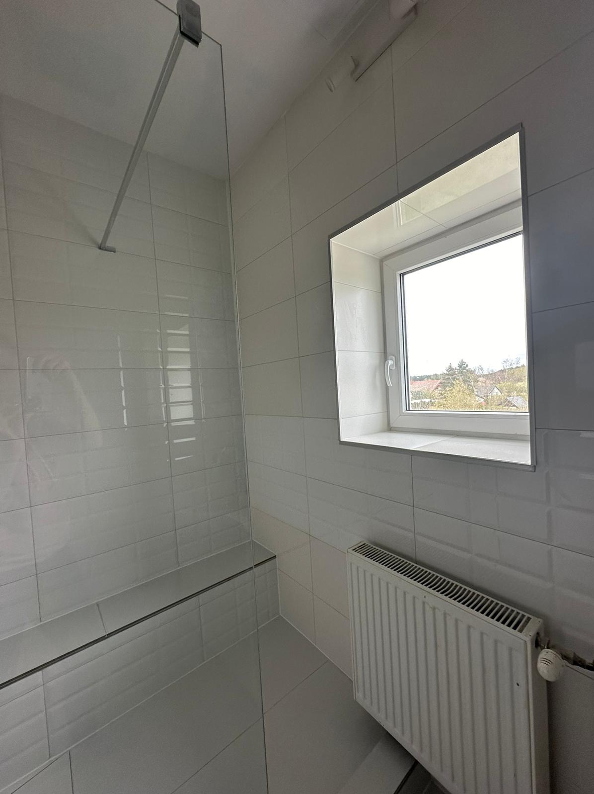 Wykończona łazienka w jasnych odcieniach szarości z kabiną prysznicową typu walk-in, grzejnikiem żeberkowym pod oknem i płytkami imitującymi cegłę.