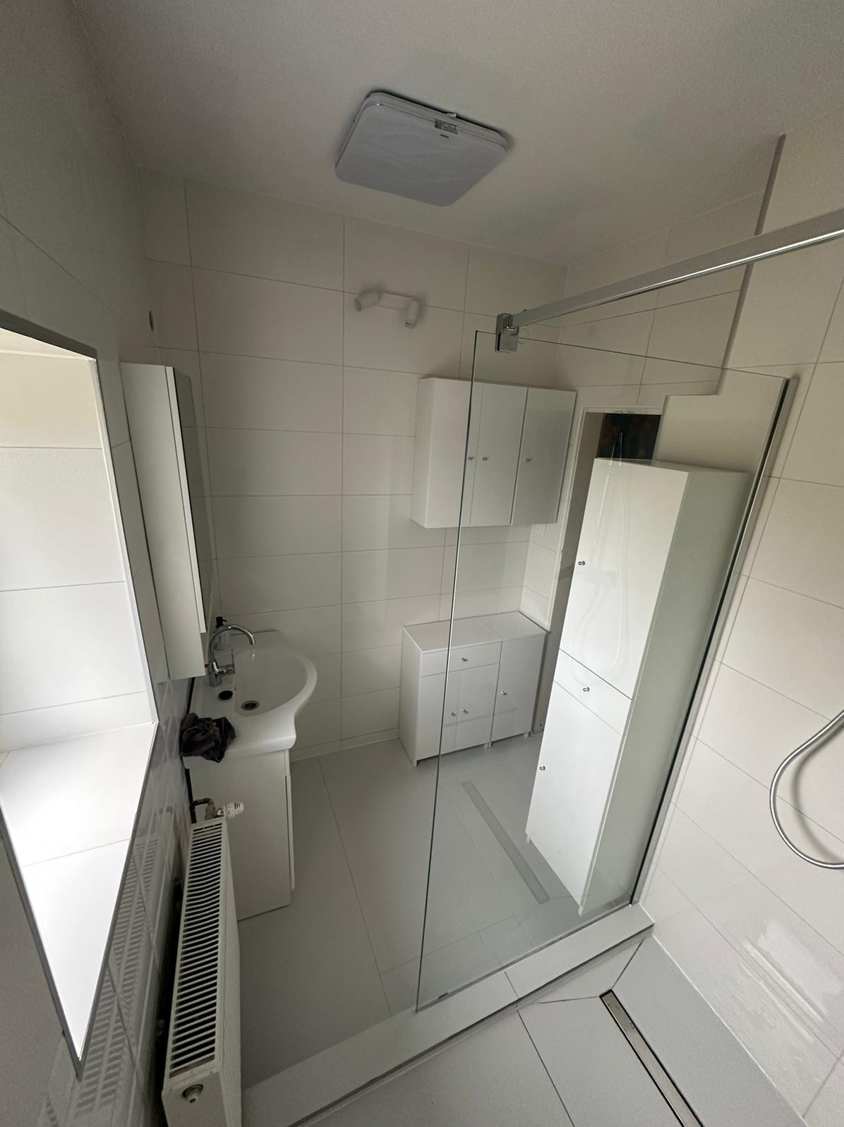 Jasna łazienka z białymi płytkami, umywalką na postumencie, grzejnikiem żeberkowym, szafkami wiszącymi i stojącymi oraz kabiną prysznicową ze szklaną ścianką.