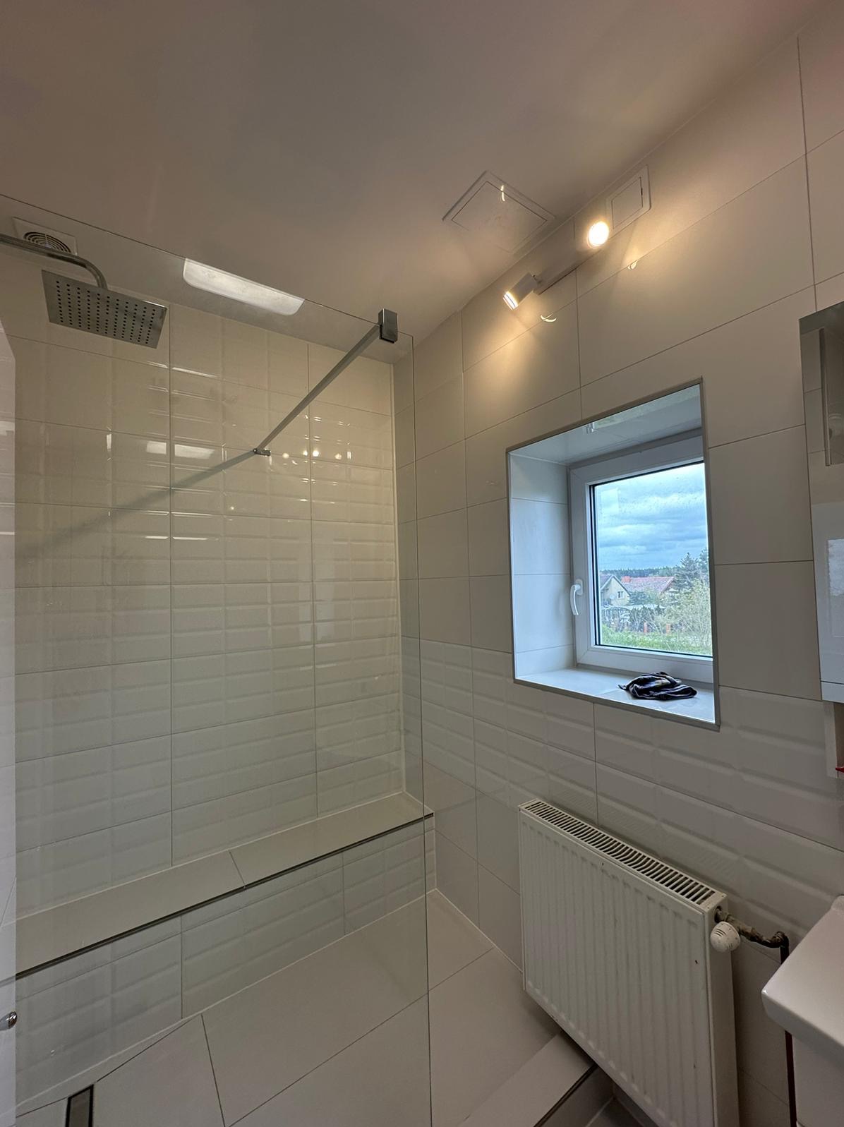 Nowoczesna kabina prysznicowa z białymi, podłużnymi płytkami, przeszkloną ścianą i siedziskiem, widok na okno z parapetem i grzejnik.